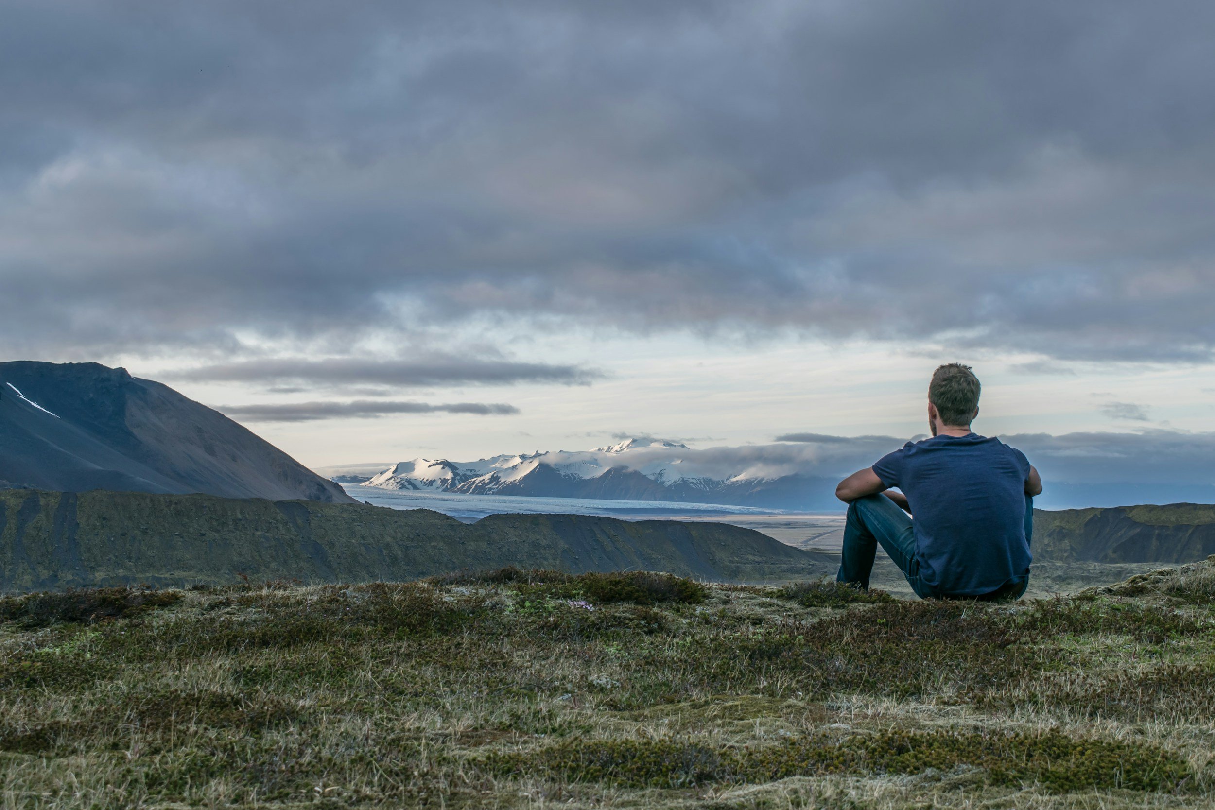 A man sitting on a grassy hill, gazing at a scenic view of snow-capped mountains, a valley, and cloudy sky.