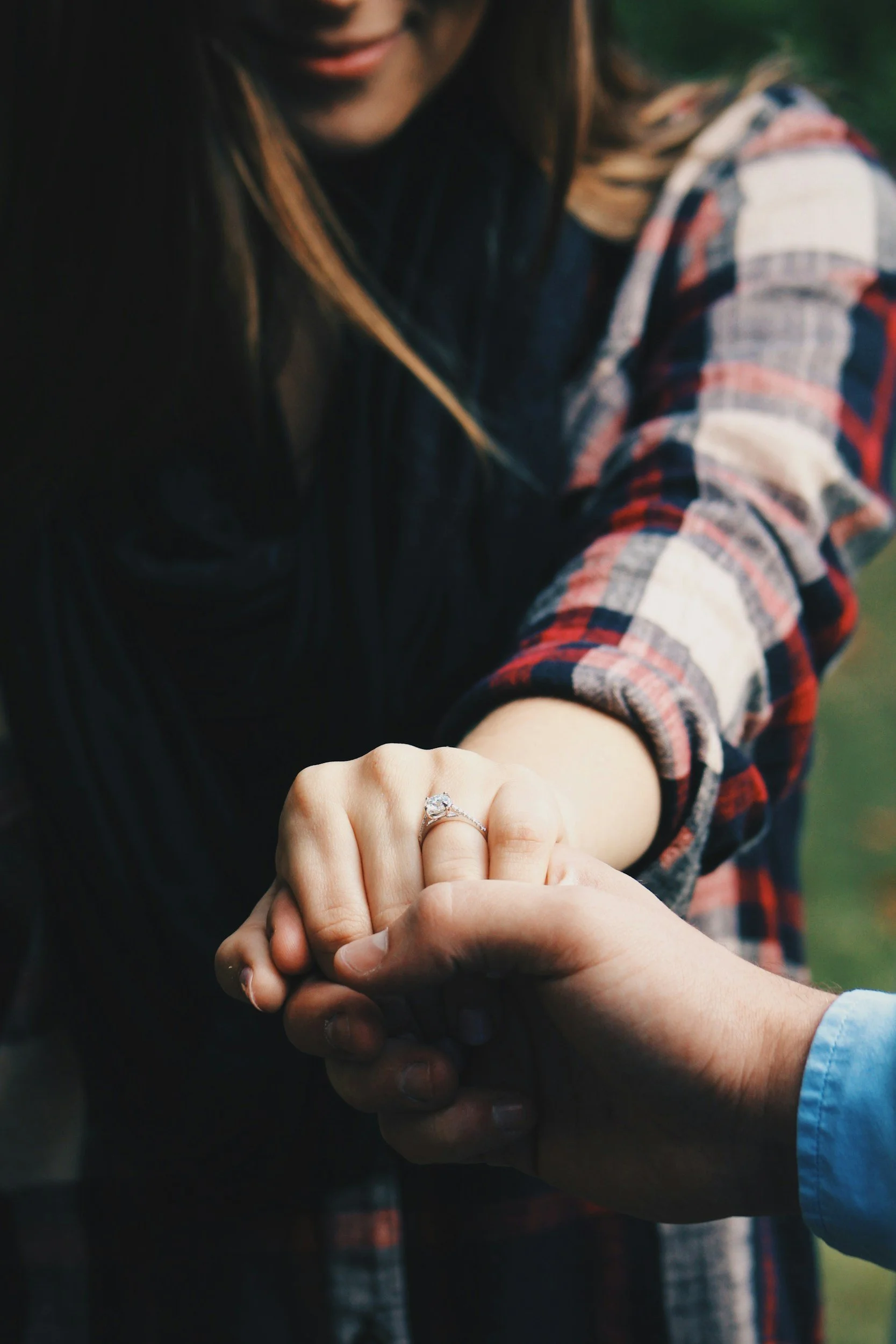 A woman showing her left hand with an engagement ring, holding hands with a man wearing a light blue shirt.