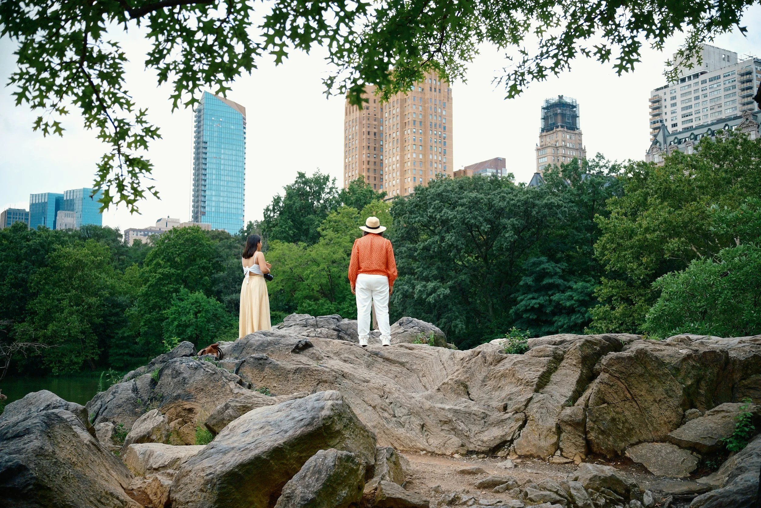 Two women standing on rocky terrain in a park with tall city buildings in the background.