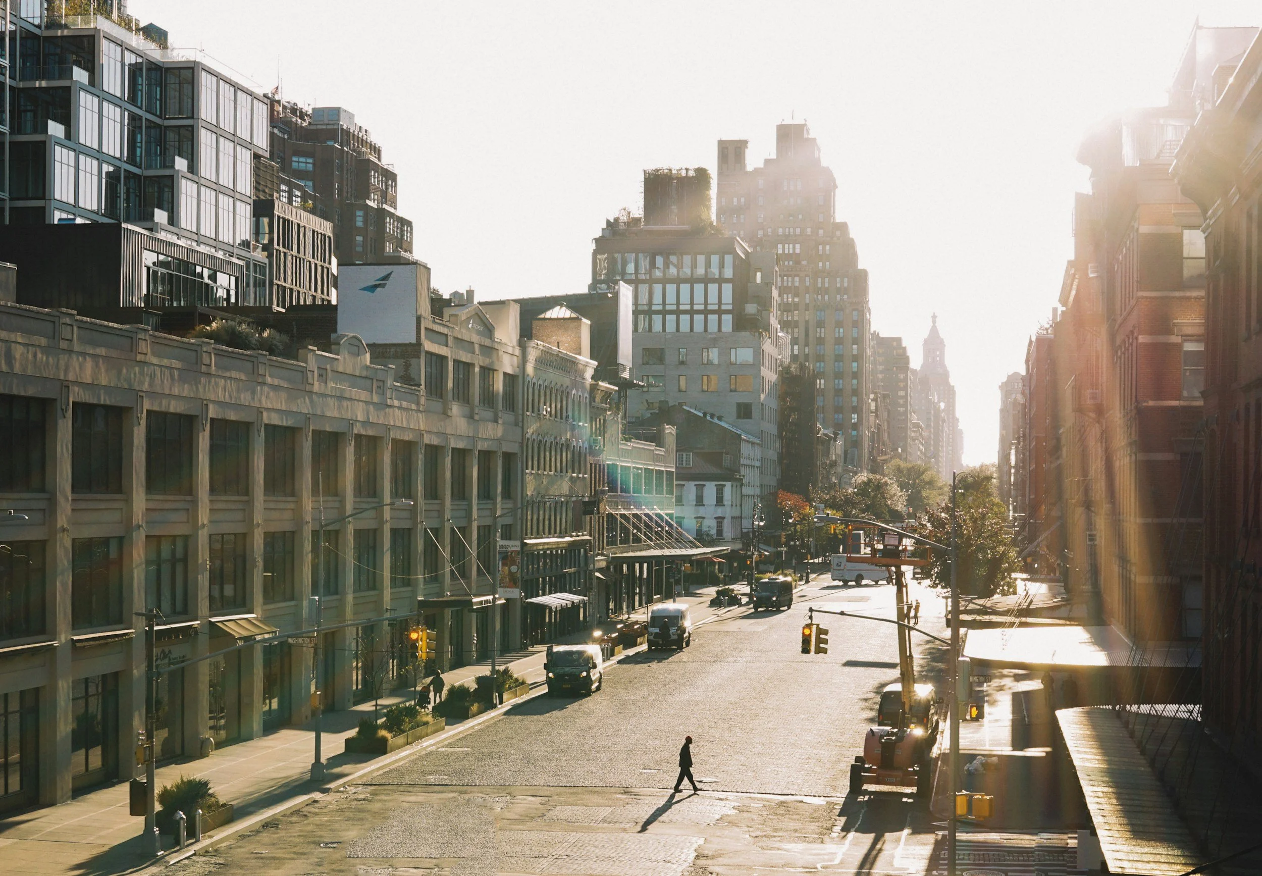 A city street scene in morning sunlight with a single pedestrian crossing the road, surrounded by modern and historic buildings, cars parked along the street, and a yellow traffic light.