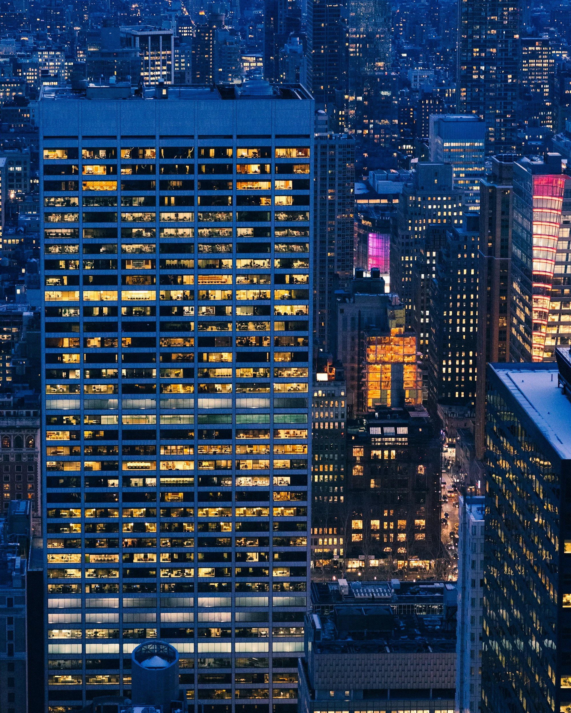 Nighttime cityscape of tall buildings with illuminated windows in a downtown area.
