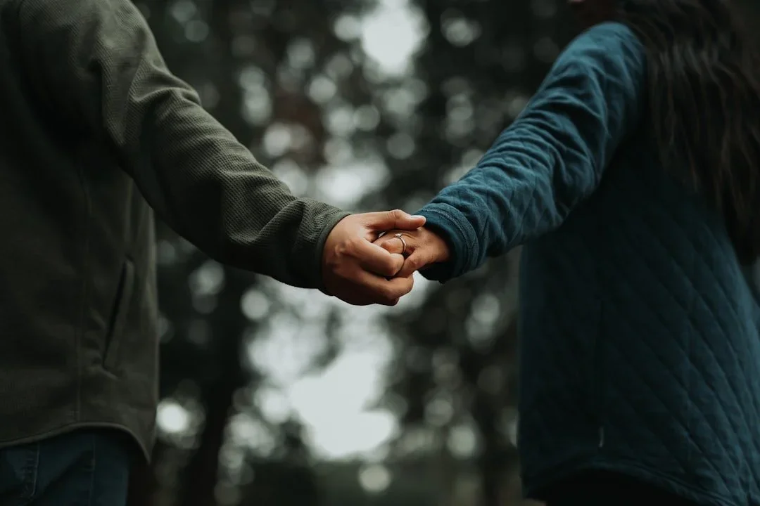 Two people holding hands outdoors, with one person wearing a dark jacket and the other wearing a blue quilted jacket.