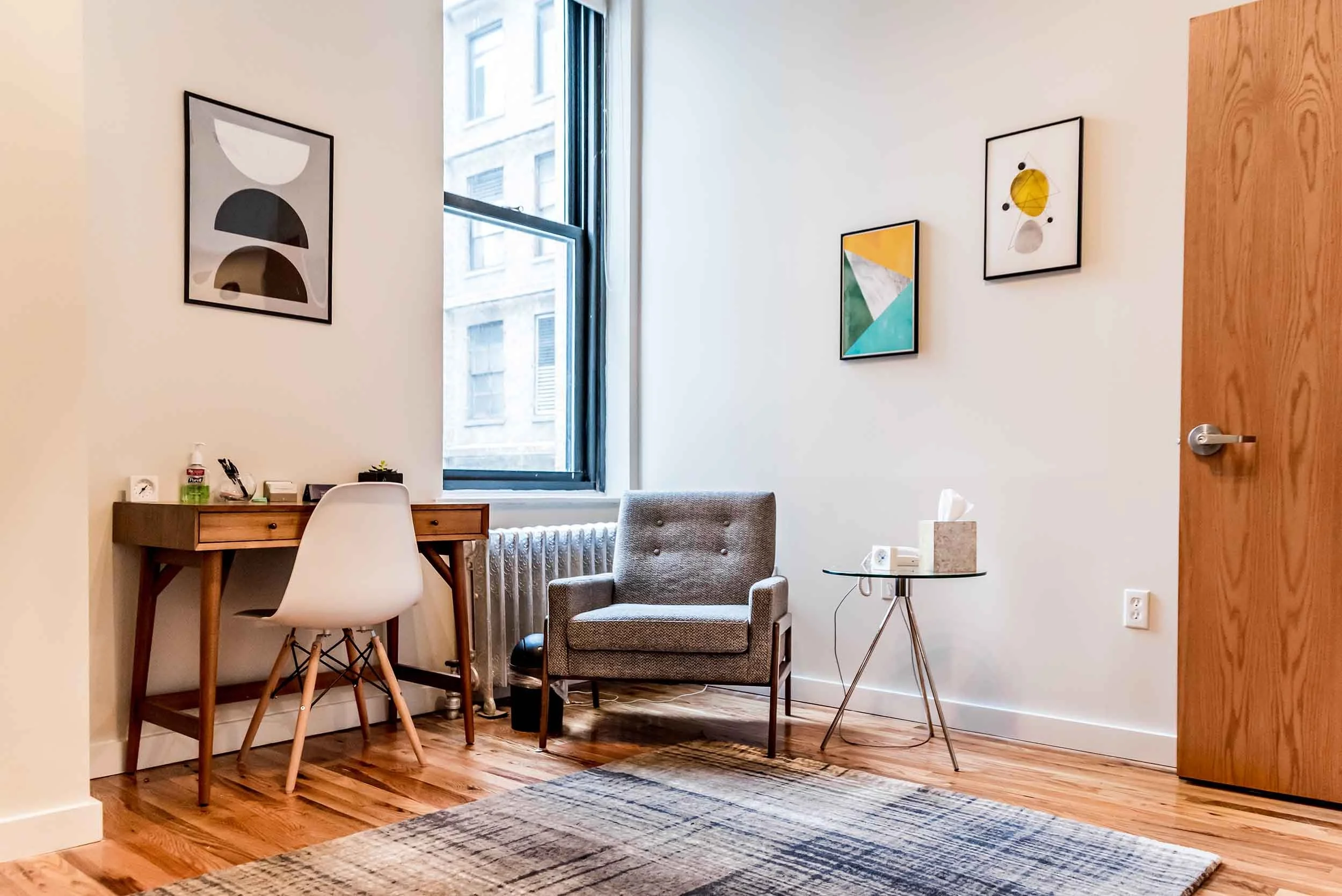 Minimalist room with a desk and chair, a gray armchair near a window, a small side table with a tissue box and phone, artwork on the white walls, hardwood floor, and a patterned rug.