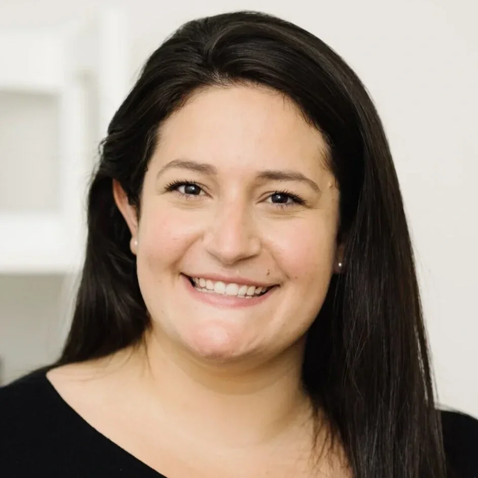 Close-up portrait of a woman with long dark hair, smiling, wearing earrings and a black top, against a neutral background.