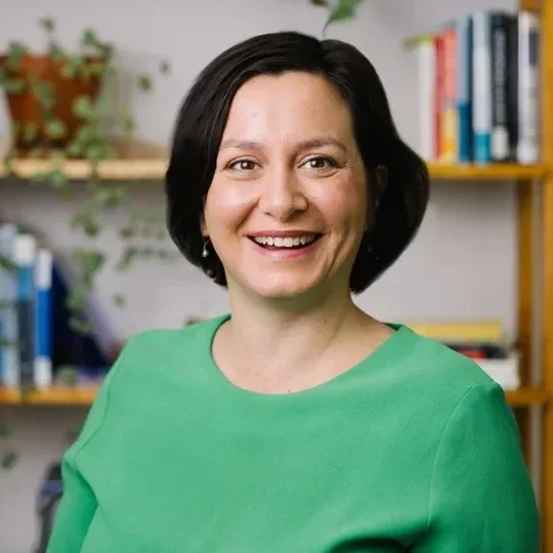 A smiling woman with black hair, wearing a green top, standing indoors in front of a bookshelf with books and plants.