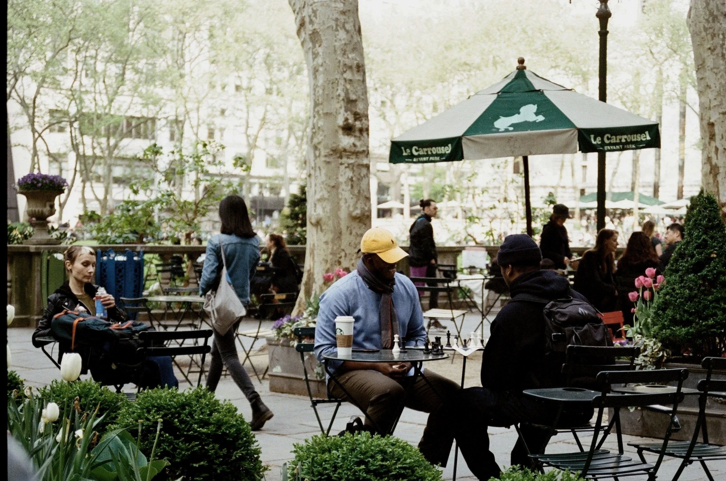 People sitting at tables in a park with trees and flowers, including tulips, some people walking, and an outdoor cafe with an umbrella that reads "Le Carrousel."
