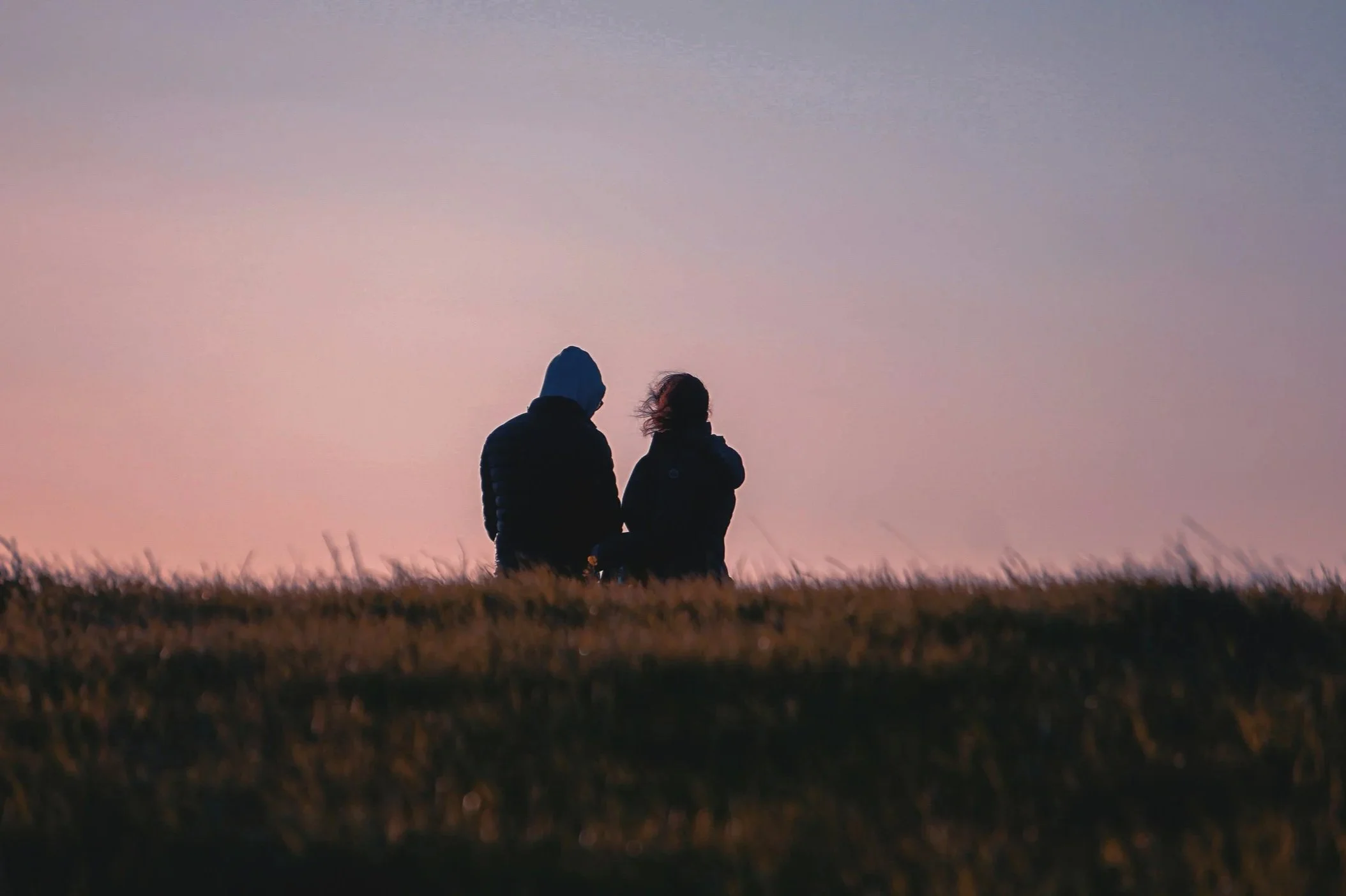 Two people sitting on a grassy hill at sunset or sunrise, facing away from the camera, with a pastel-colored sky.