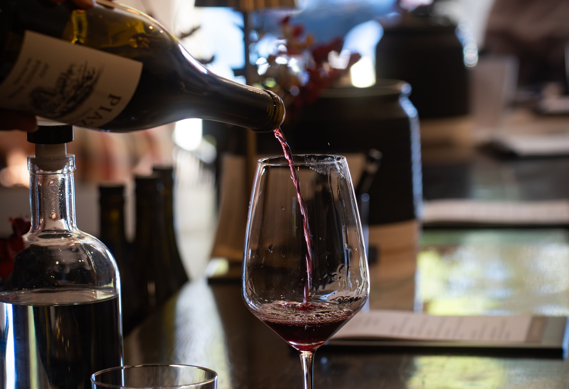 Pouring red wine into a wine glass in a cozy bar or restaurant setting with bottles and decorative objects in the background.