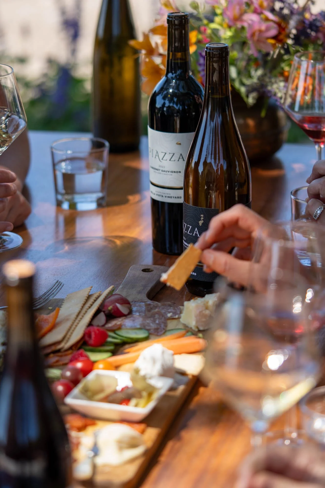 A cheese and charcuterie tasting with wine bottles, glasses, and a cheese board featuring cheese, vegetables, grapes, and meats on a wooden table with a vase of flowers in the background.