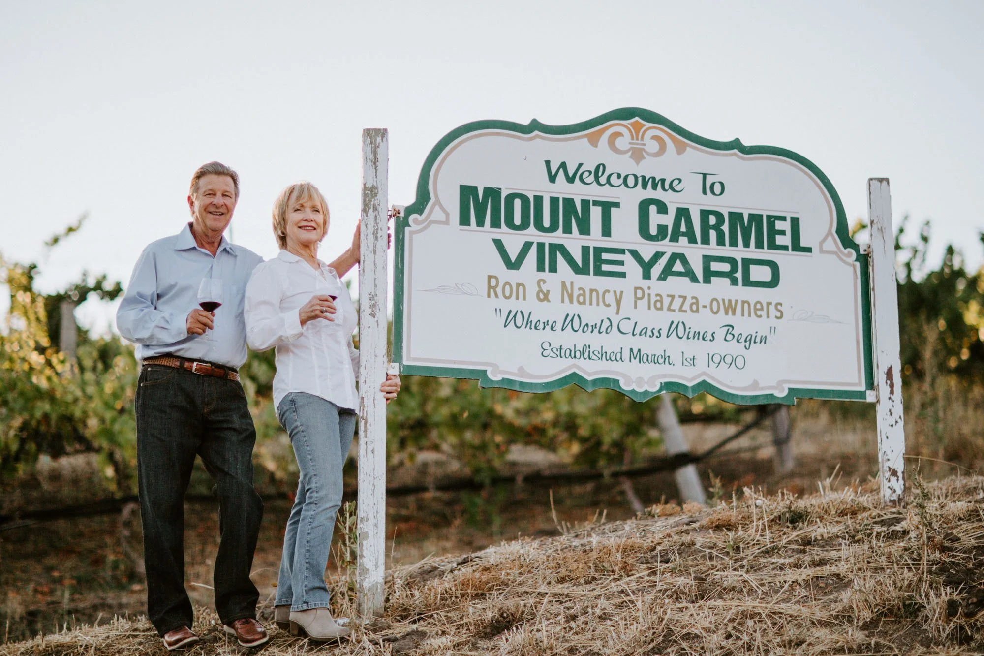 A happy couple standing outdoors next to a sign that says "Welcome to Mount Carmel Vineyard"; they are holding glasses of red wine and smiling, with vineyards in the background.