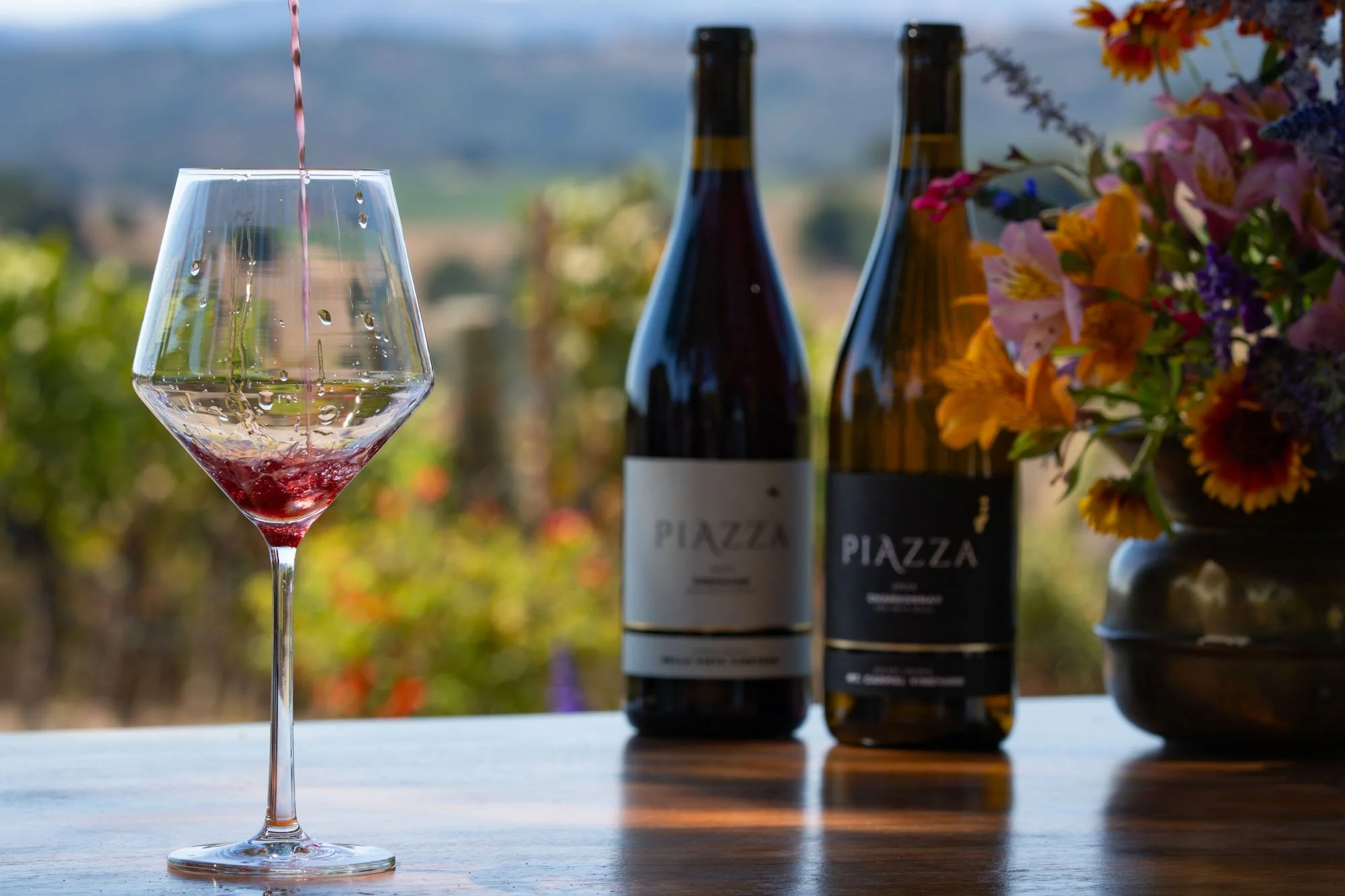 A glass of red wine being poured with a vineyard in the background, two bottles of Piazzo wine on the table, and a vase of colorful flowers.