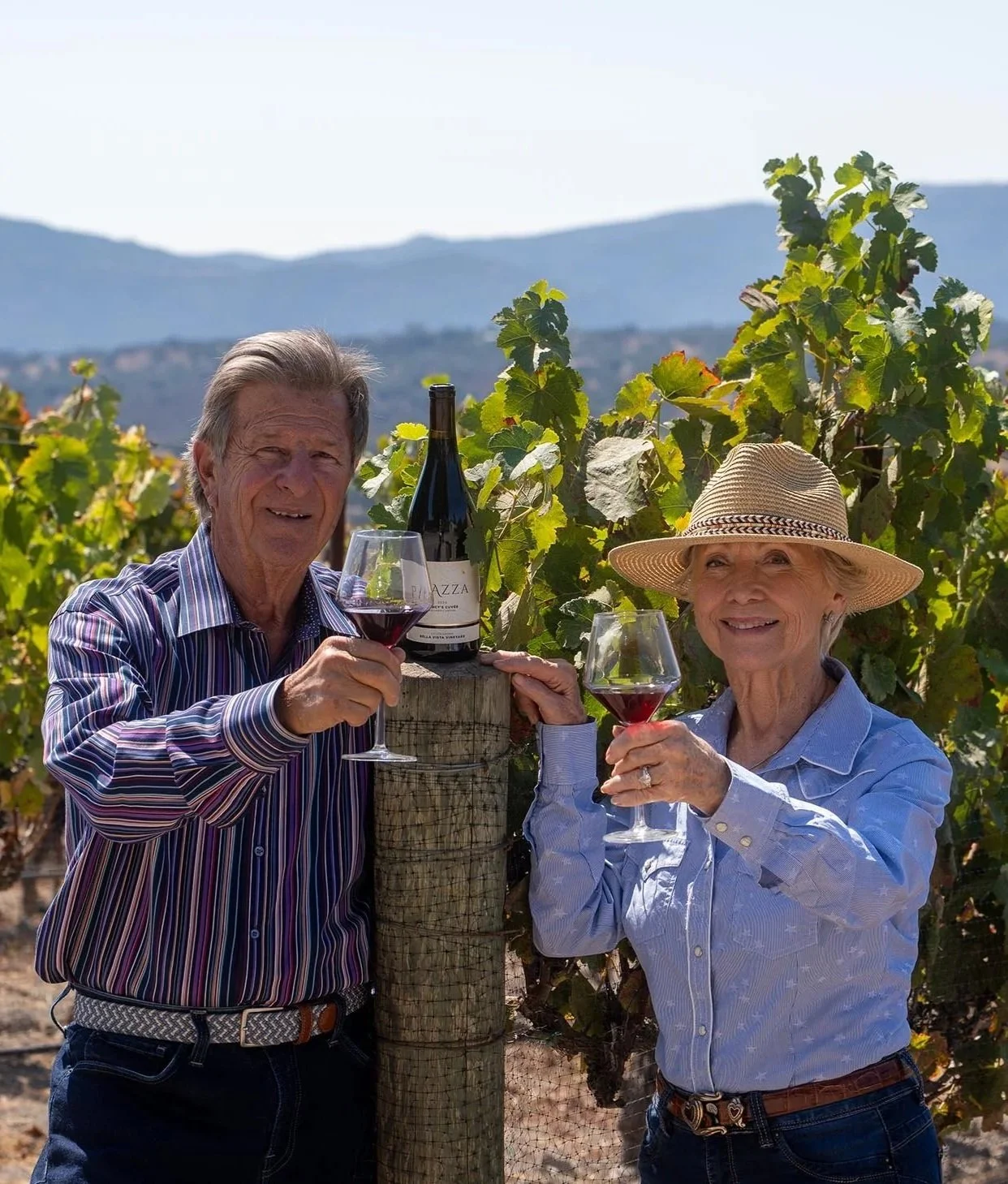 A man and woman standing in a vineyard, holding glasses of red wine, smiling, with a bottle of wine on a wooden post between them and mountains in the background.
