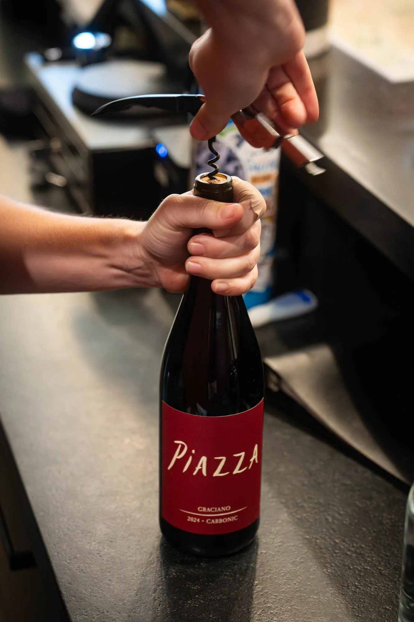 Person opening a brown glass bottle of Piazza Craciano 2021 Carbonic wine with a corkscrew in a kitchen or bar setting.