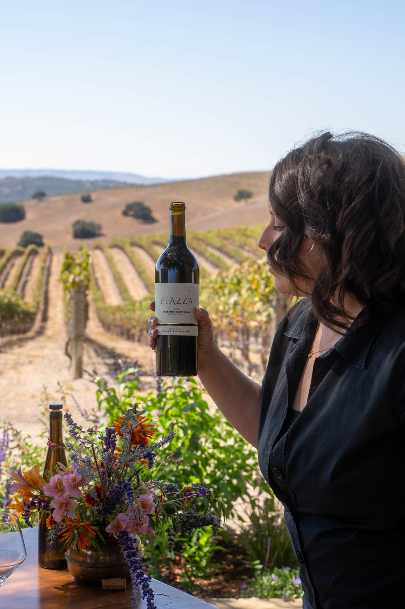 A woman with dark, wavy hair in a black shirt holding a bottle of Piazzza Cabernet Sauvignon wine, with a vineyard and rolling hills in the background.