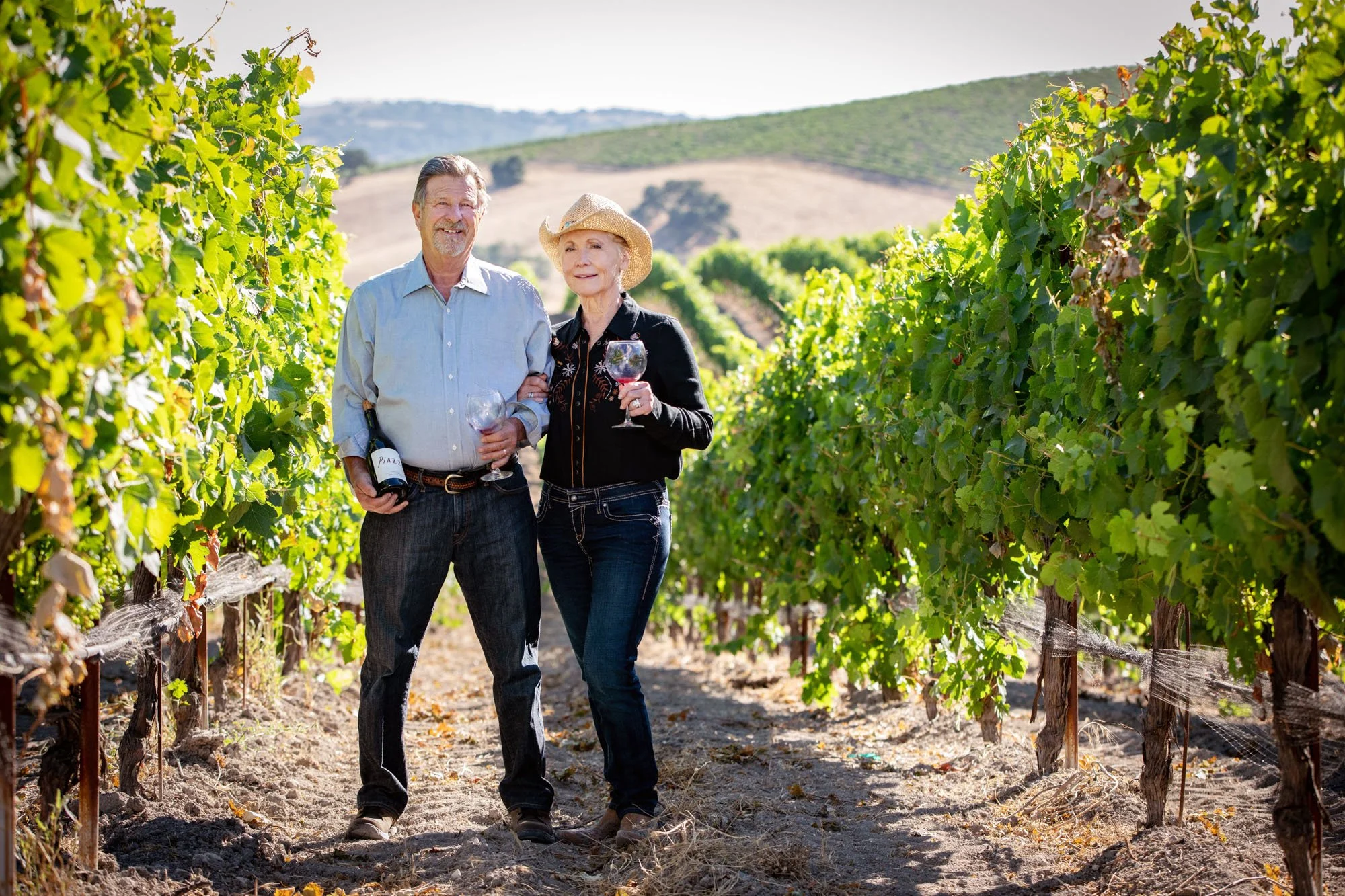 A man and woman stand together in a vineyard, smiling and holding glasses of red wine. The man also holds a bottle of wine. The vineyard has lush green grapevines, and rolling hills can be seen in the background on a sunny day.