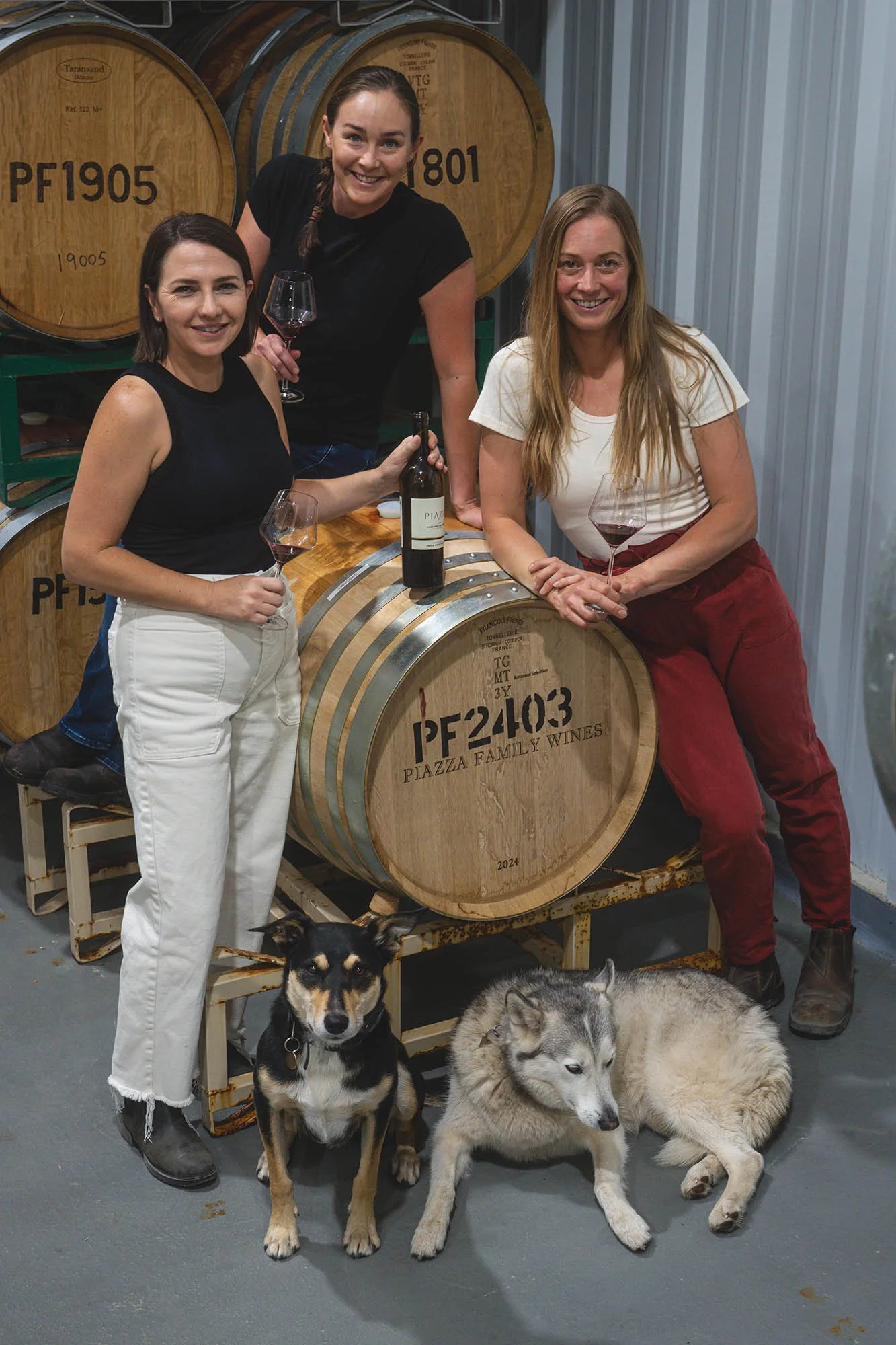 Three women holding glasses of red wine and a bottle of wine in a winery with large wine barrels in the background, and two dogs sitting on the floor nearby.