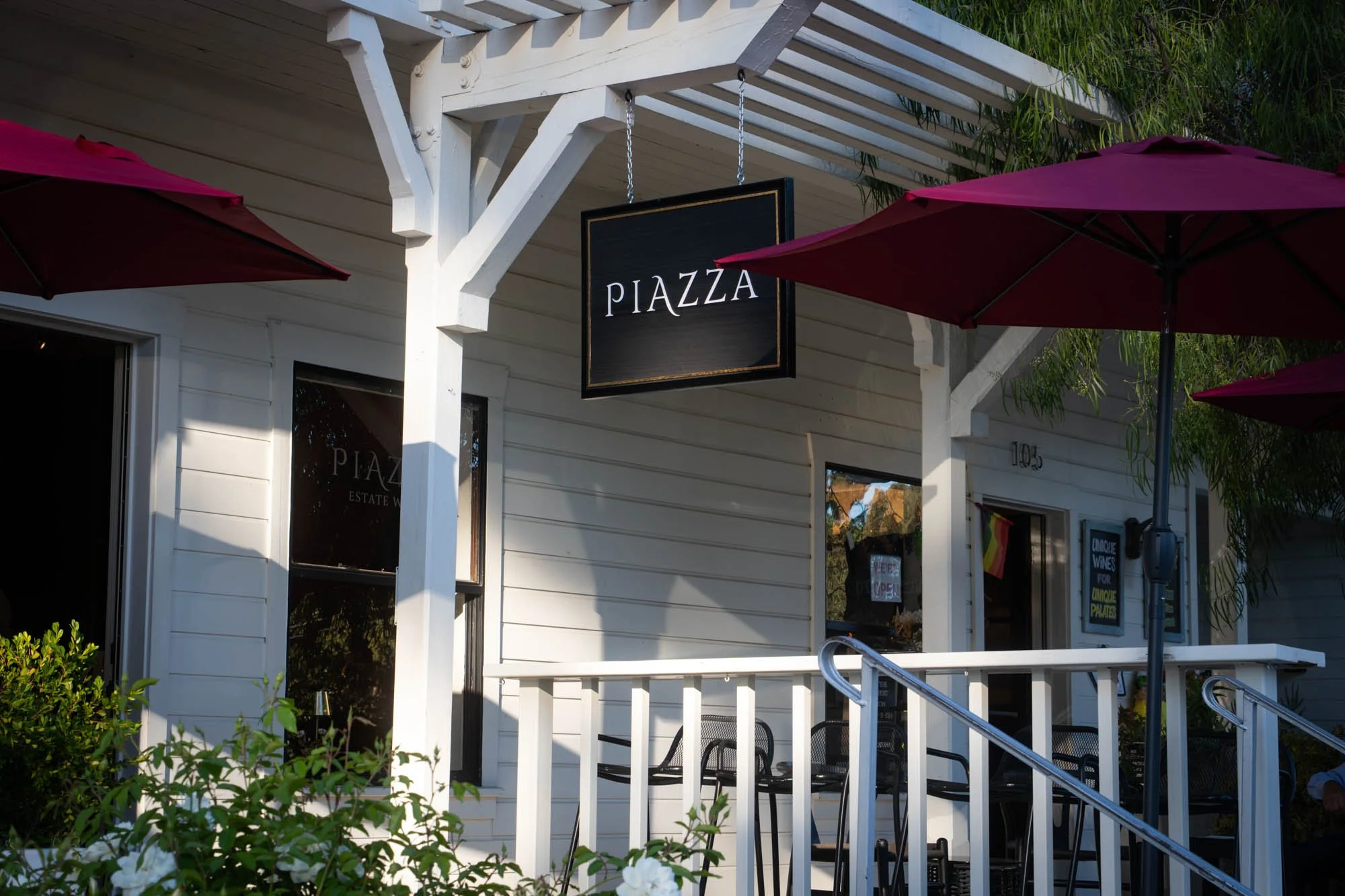 Exterior of a white building with a black sign that reads 'PIAZZA' hanging above the porch. The porch has a white railing, black chairs, and large pink umbrellas providing shade. Green foliage is visible around the building.