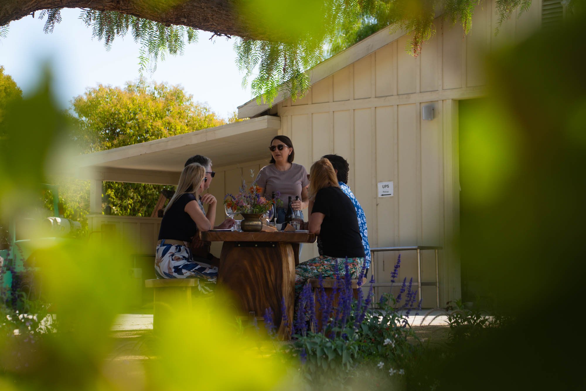 A group of five women sitting outdoors at a wooden table, engaged in conversation with one standing woman. The setting includes trees, flowers, and a small building with a UPS sign.