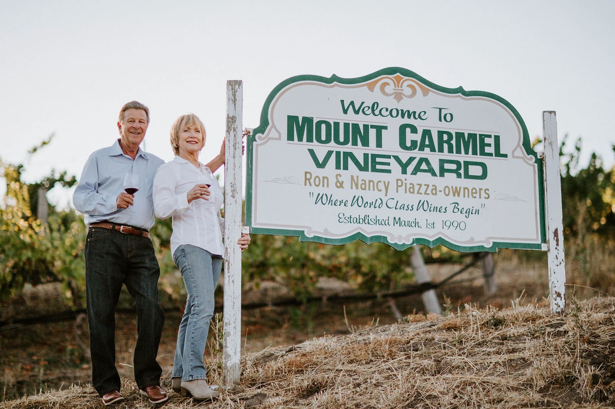 A man and woman stand beside a sign that reads "Welcome to Mount Carmel Vineyard," holding glasses of red wine and smiling. The man is dressed in a light blue shirt and dark pants, the woman in a white blouse and jeans, with a vineyard background.