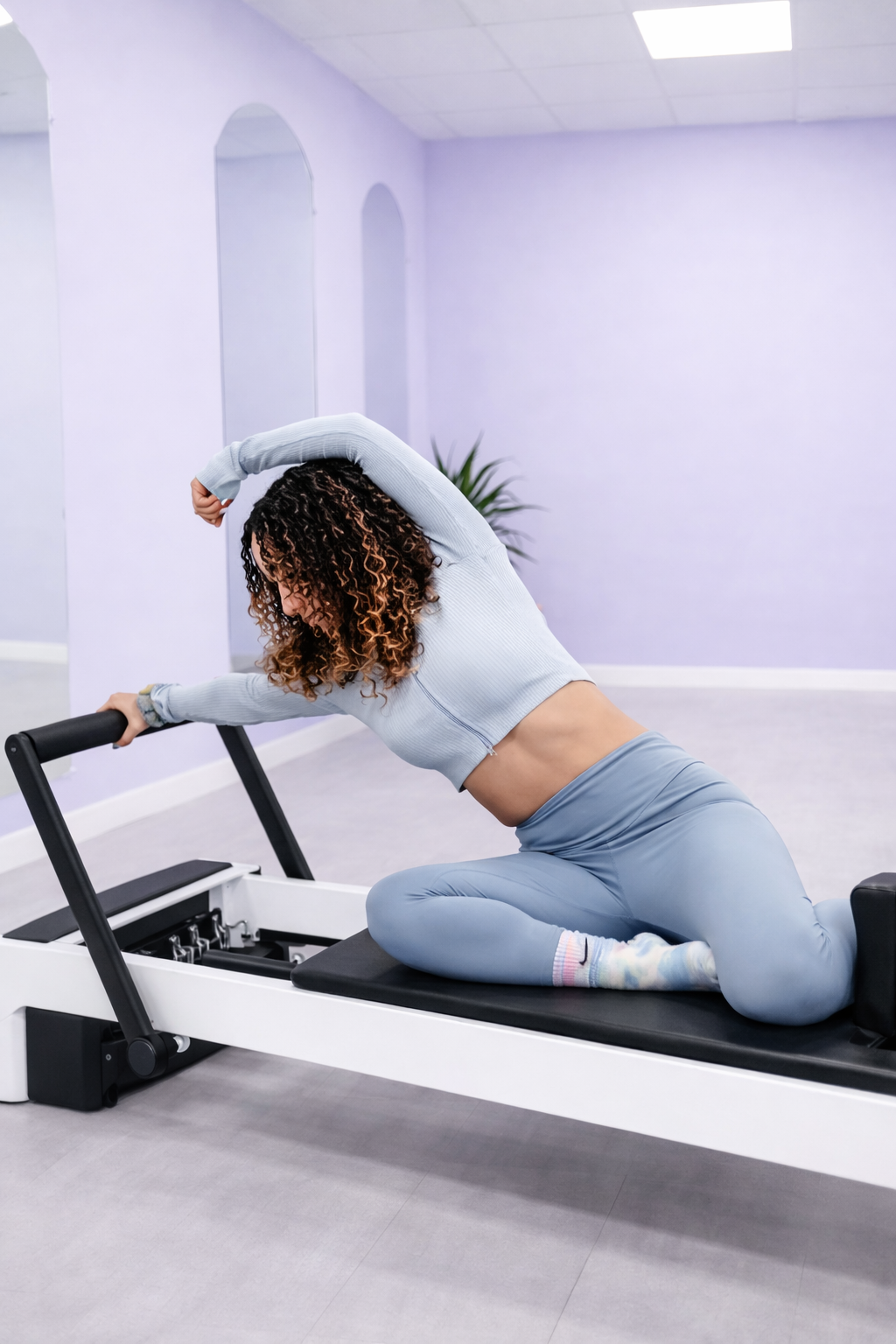 A woman with curly hair doing a workout on a Pilates reformer machine in a bright fitness studio with plants and mirrors.