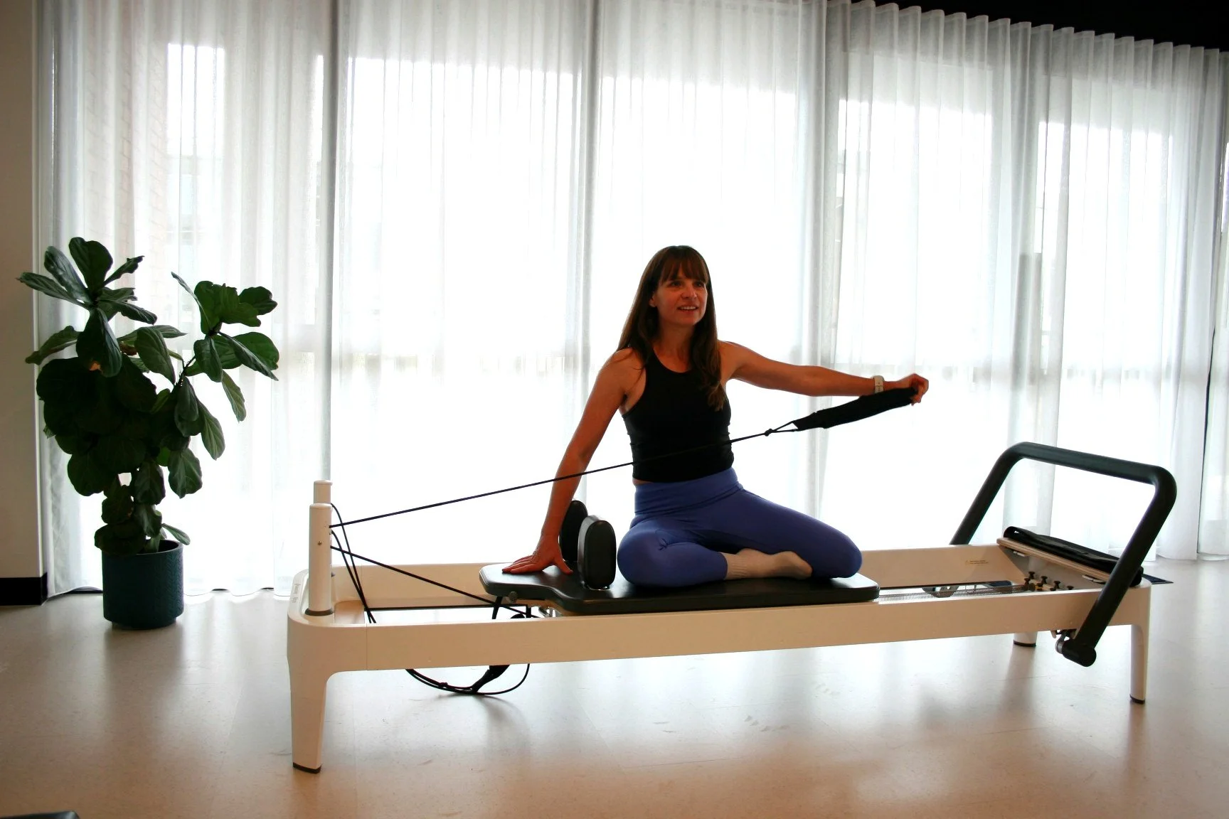 A woman practicing Pilates on a reformer machine in a bright room with sheer white curtains and a large potted plant.