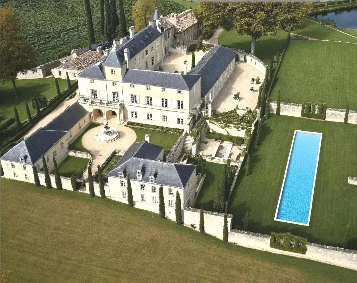 A large estate with a grand main building, fountain, swimming pool, manicured lawn, and surrounding trees and farmland.