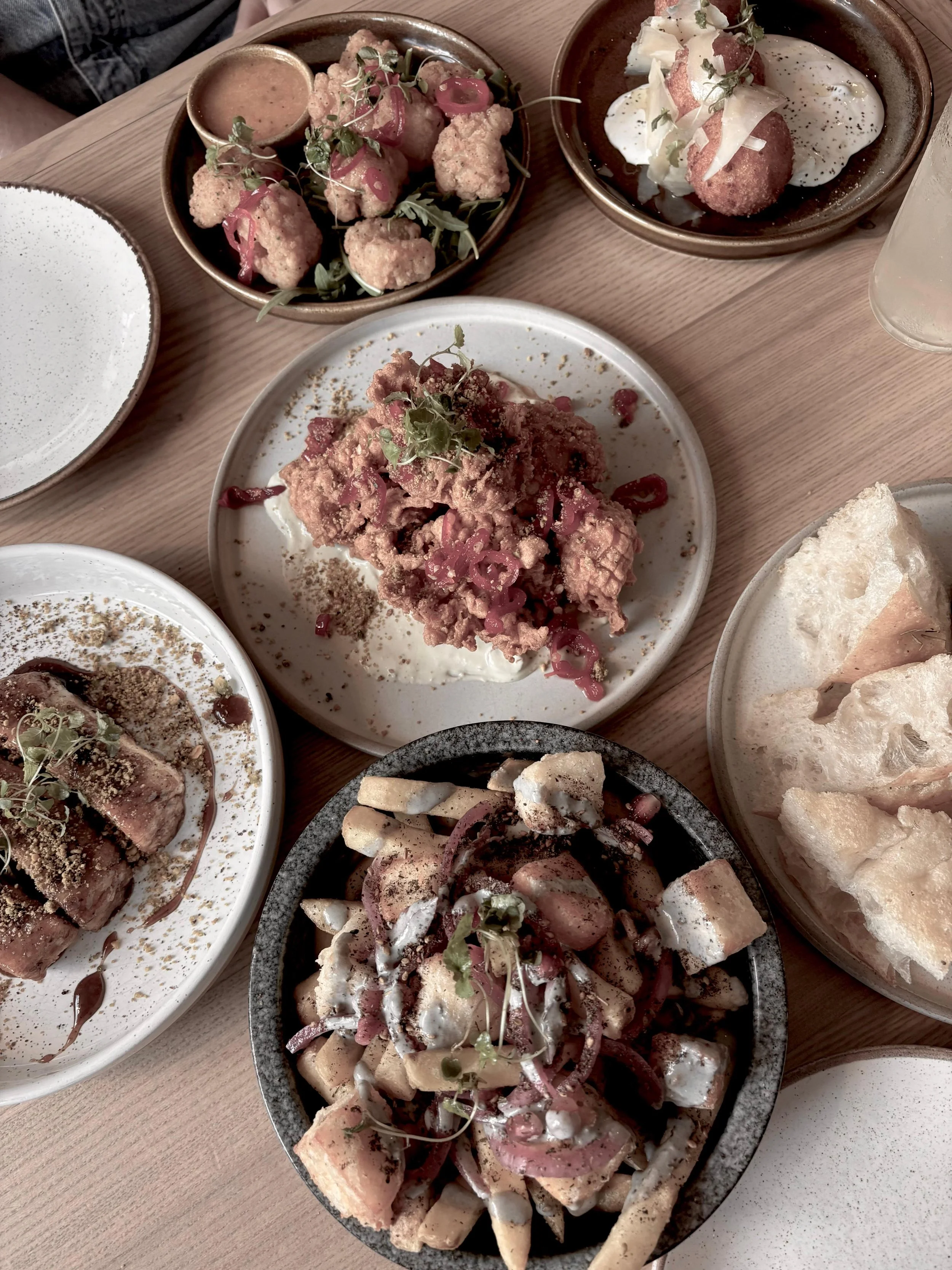 A variety of dishes on a wooden table, including a bowl of fried cauliflower, a plate of fried chicken with pink pickled onions, a bowl of salad with chopped vegetables and dressing, and slices of bread, all garnished with herbs.