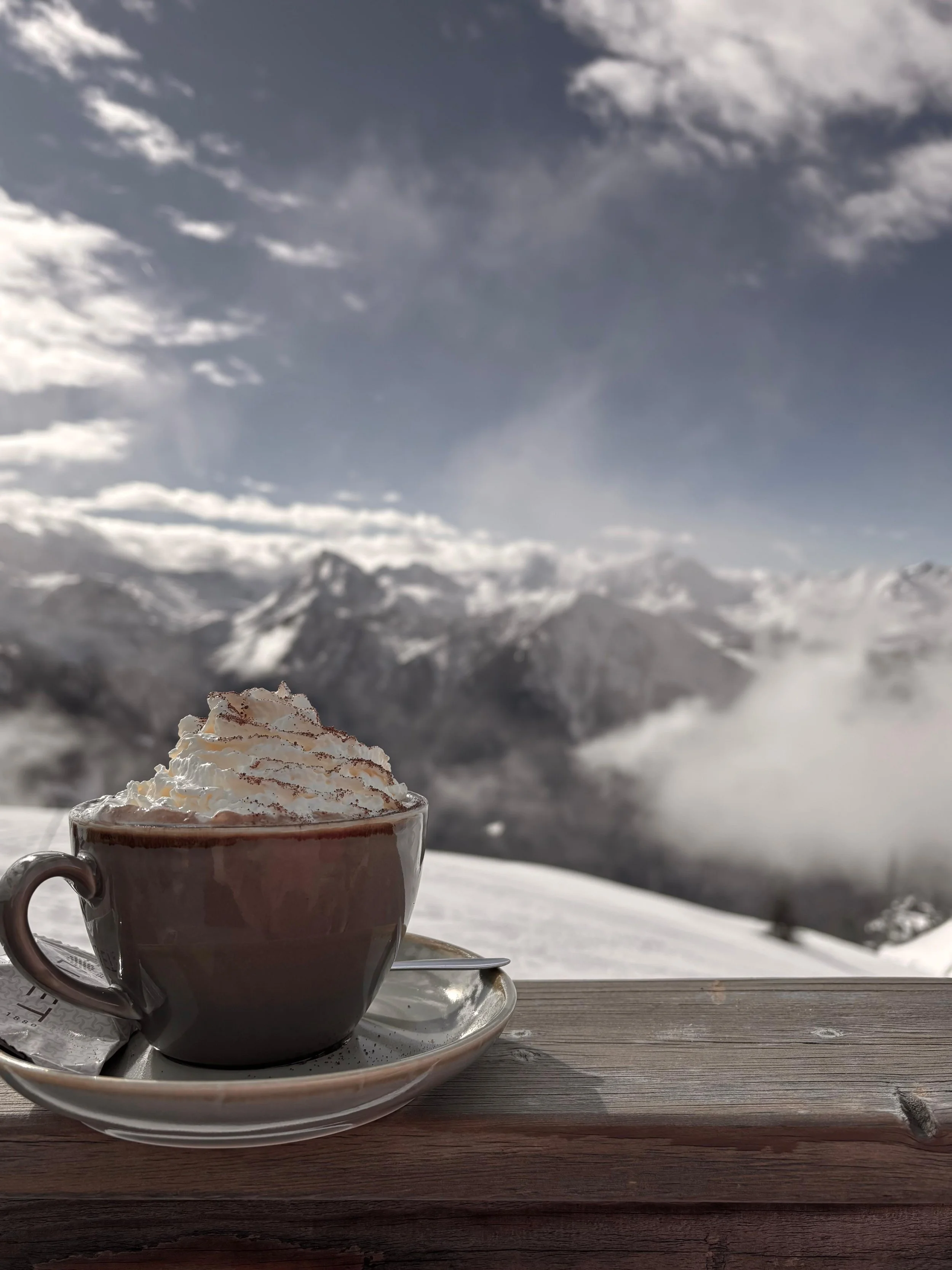A cup of hot chocolate topped with whipped cream on a saucer, placed on a wooden deck overlooking snow-covered mountains under a partly cloudy sky.