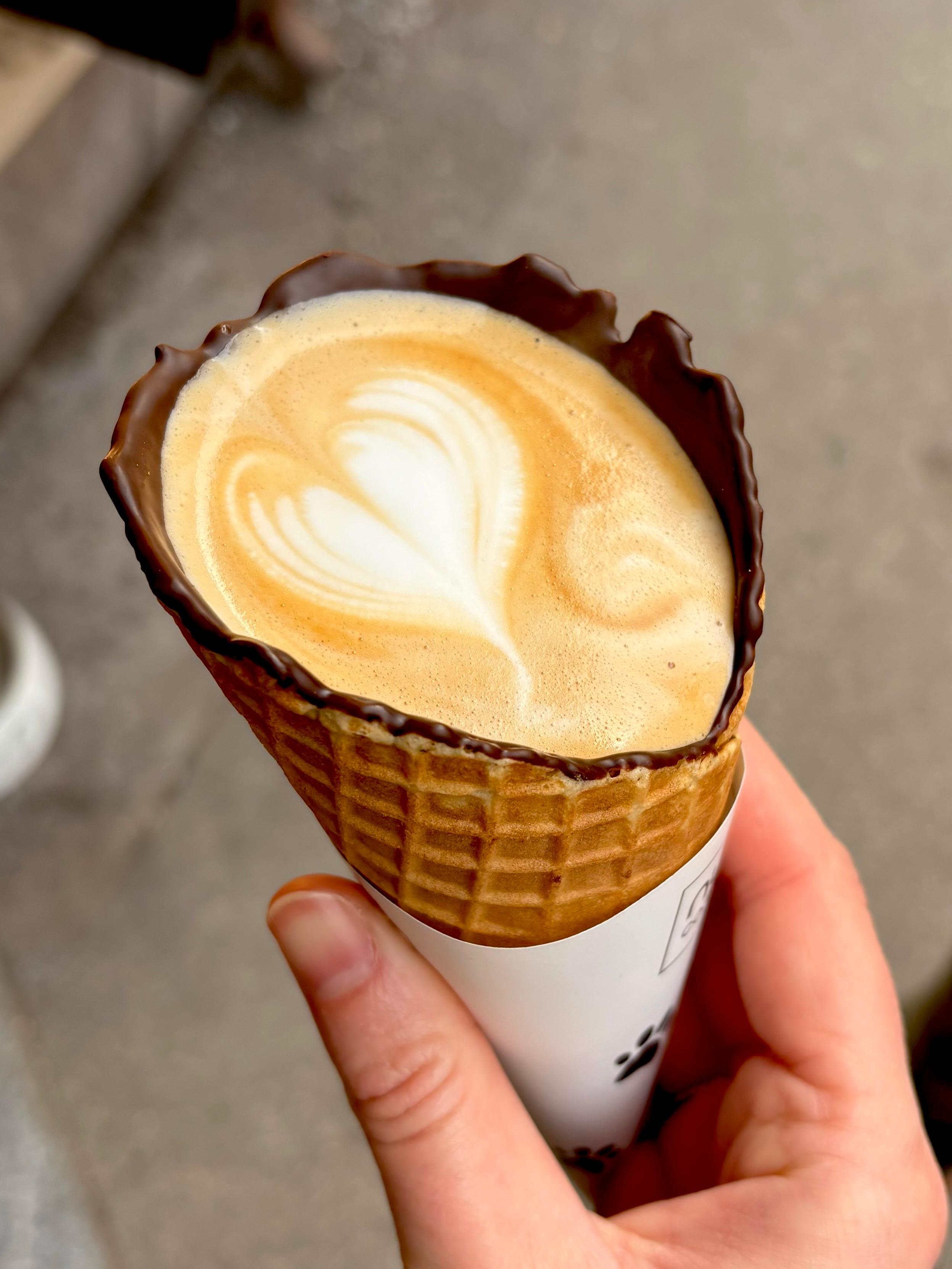 Close-up of a hand holding an ice cream cone with latte art on top, surrounded by a chocolate-dipped waffle cone.