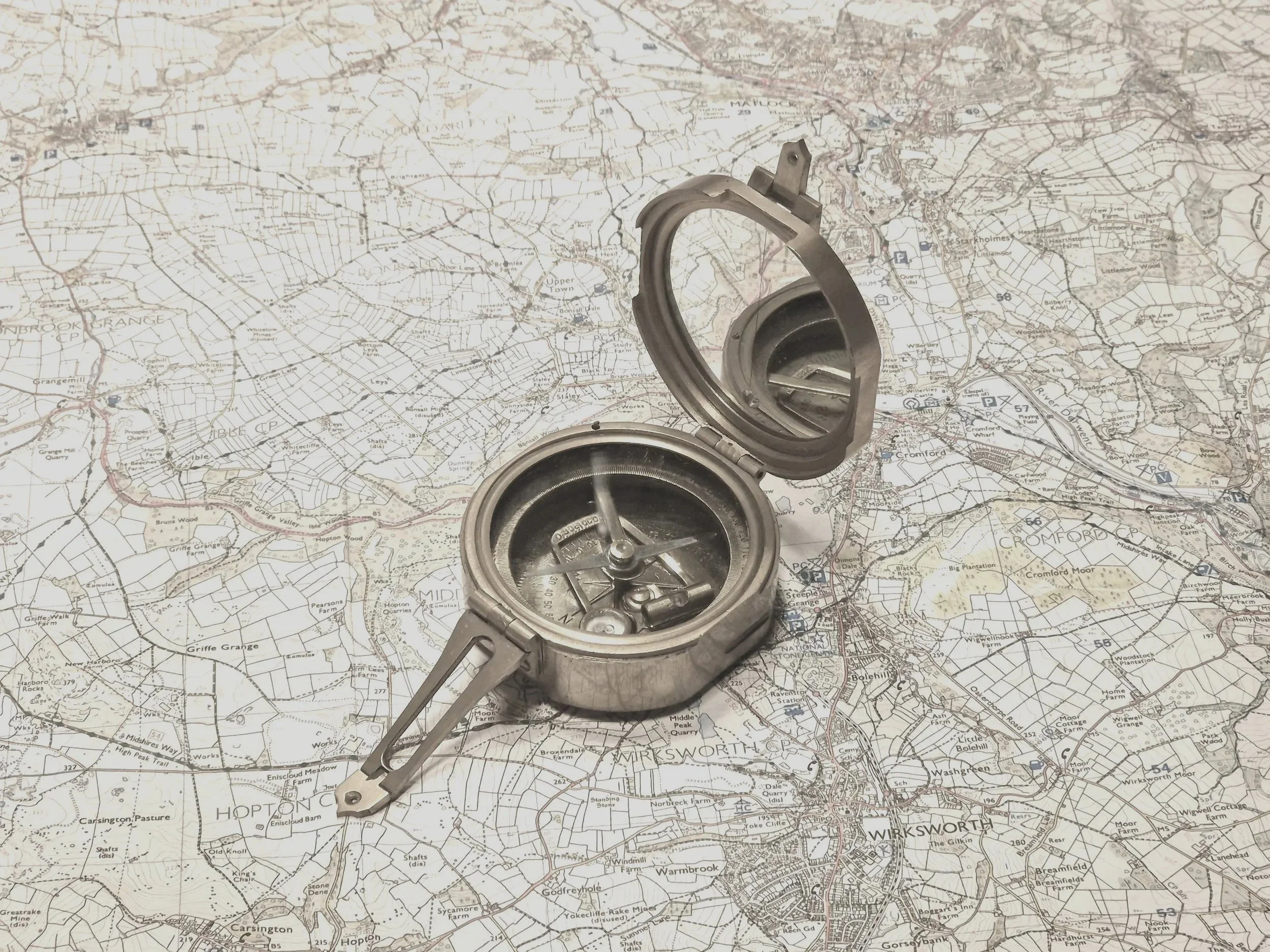 A metallic compass resting on a detailed map. The compass is open, showing its internal mechanisms and the needle pointing north.