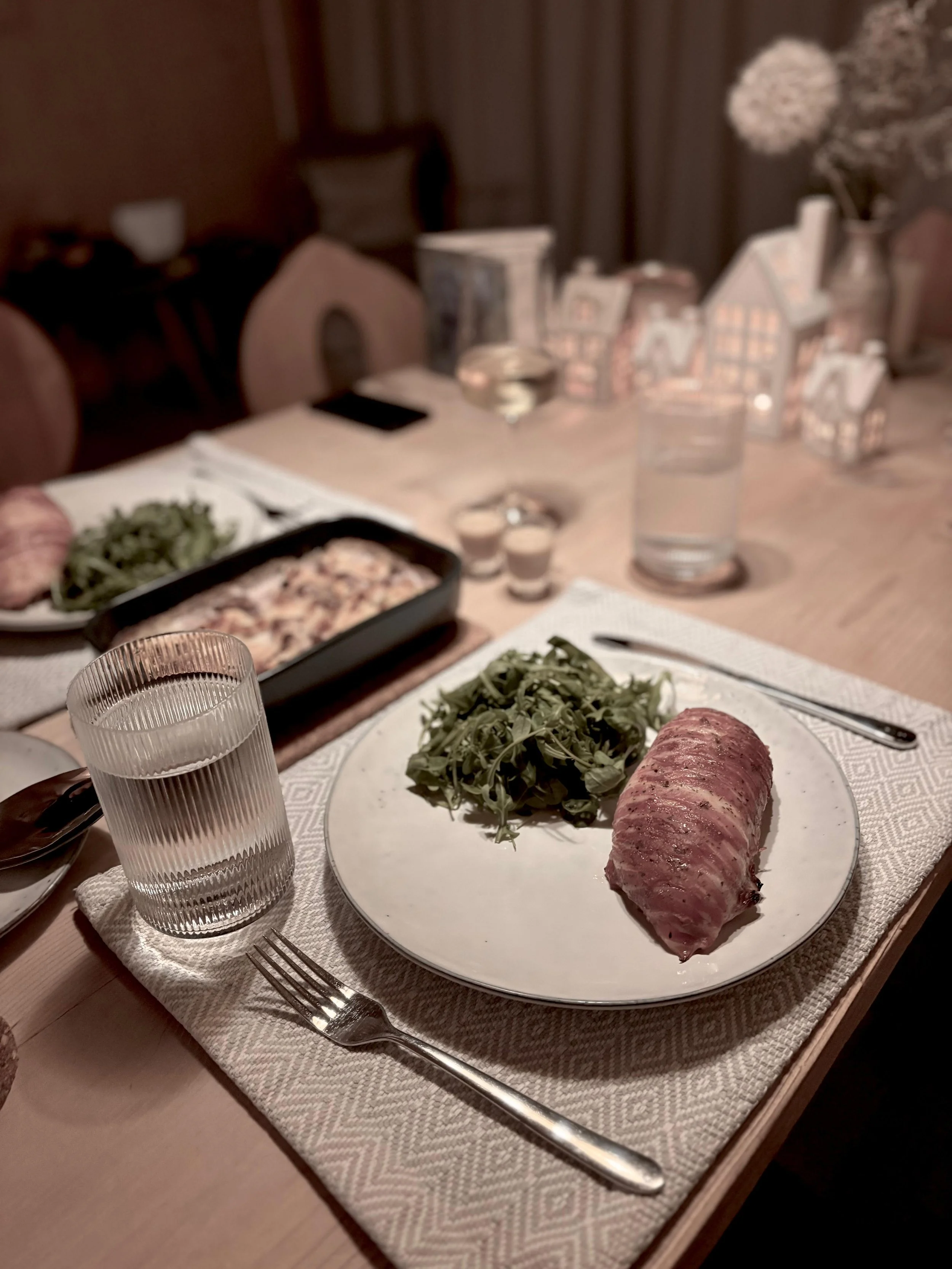 A dinner table with a plate of cooked meat and greens, a glass of water, and silverware, with a blurred background of other dishes and decor.