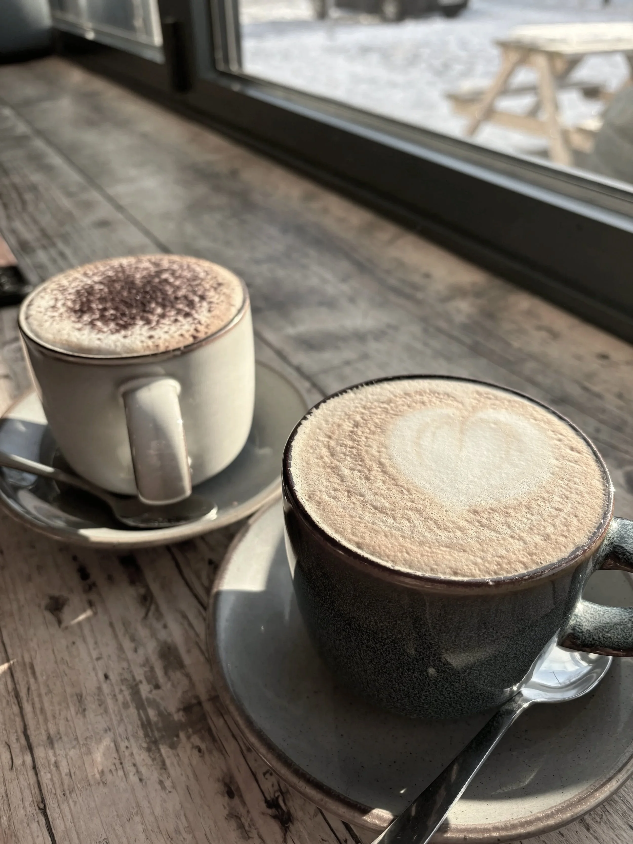Two cups of coffee with frothy milk, one with a heart-shaped latte art, placed on a rustic wooden table near a window.