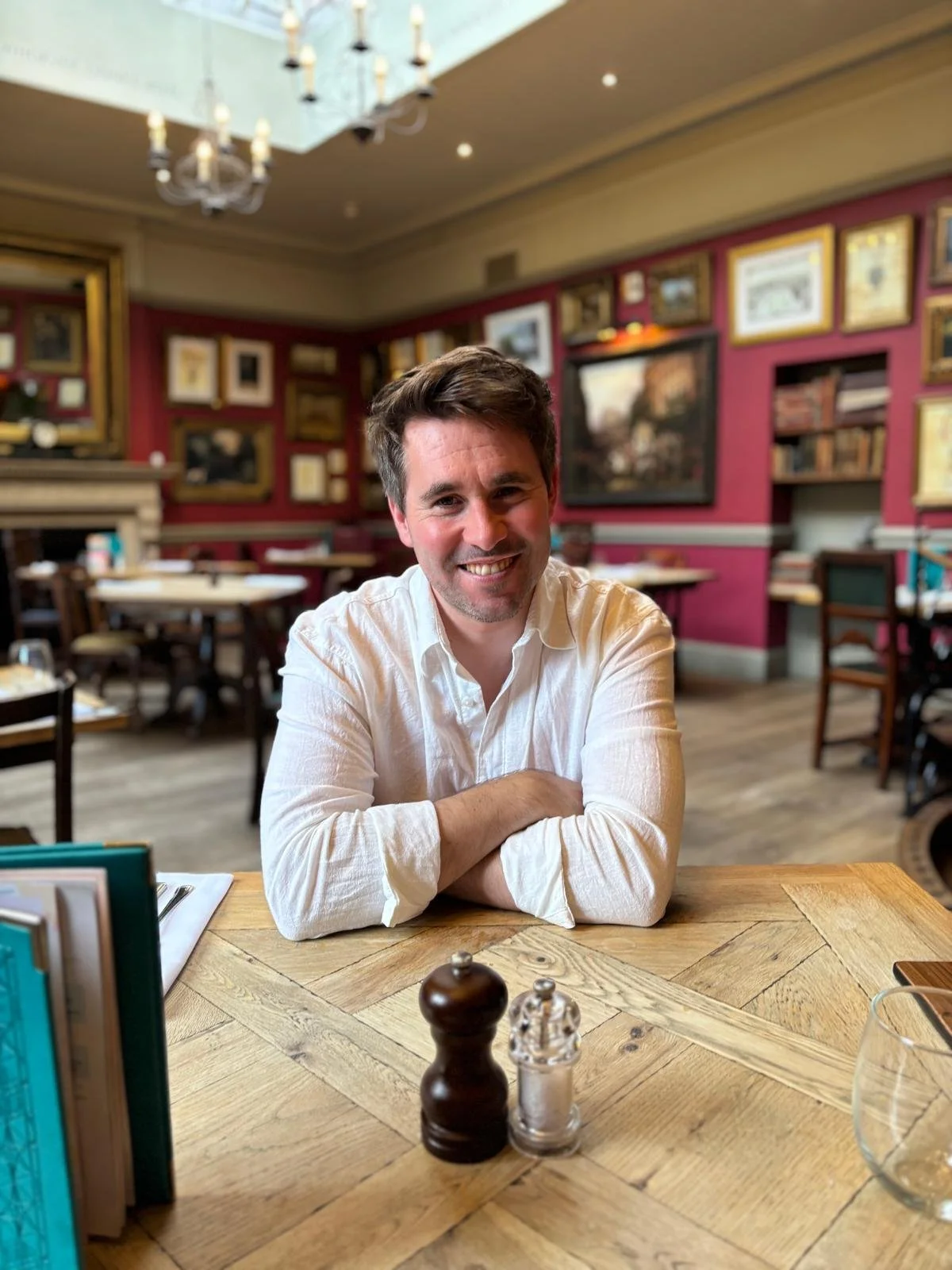 Smiling man in a white shirt sitting at a wooden table in a restaurant with framed pictures on the wall behind him.
