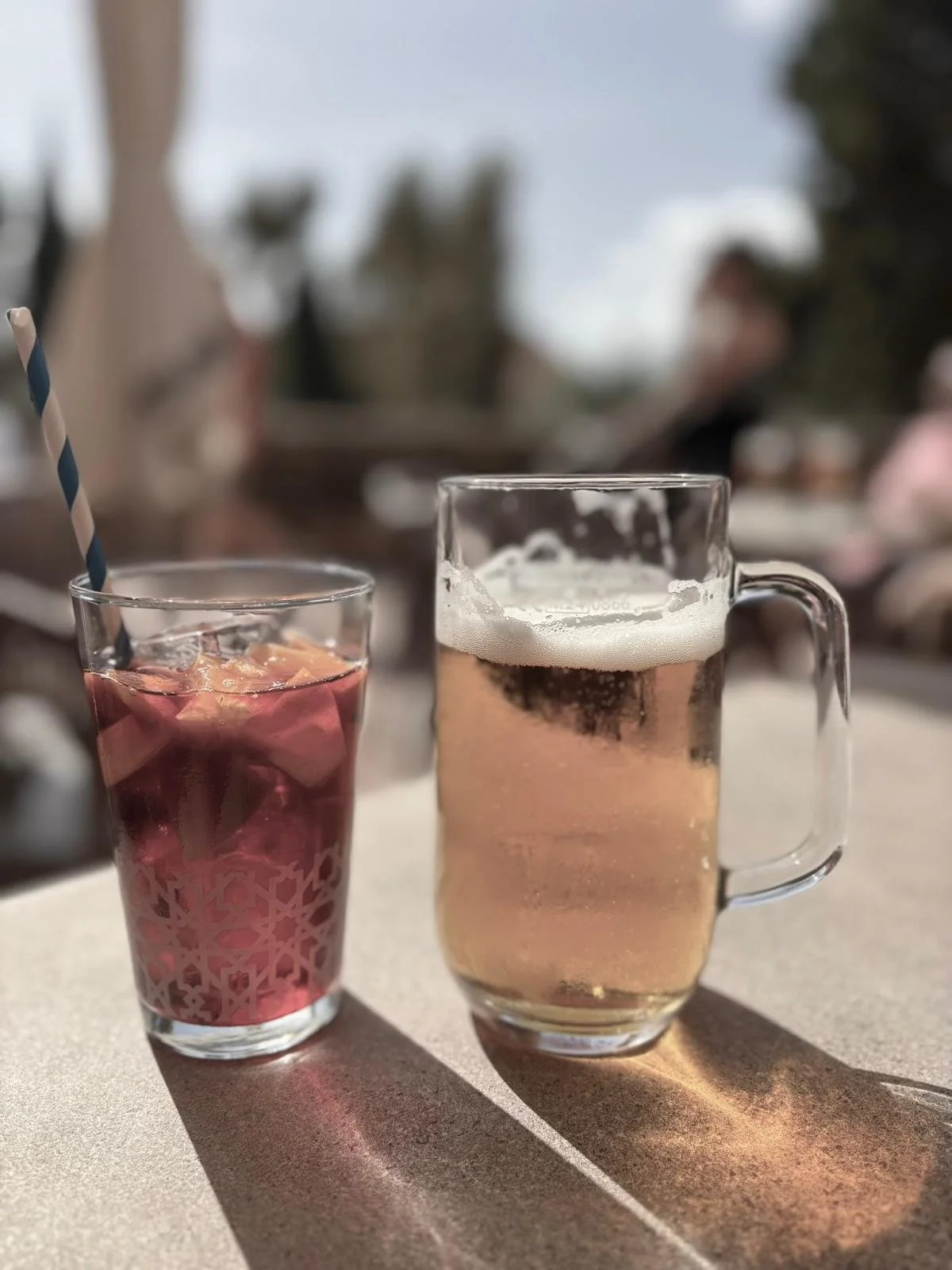 A glass of pink beverage with ice and a striped paper straw, and a mug of beer with a foamy head, on an outdoor table with sunlight casting shadows.