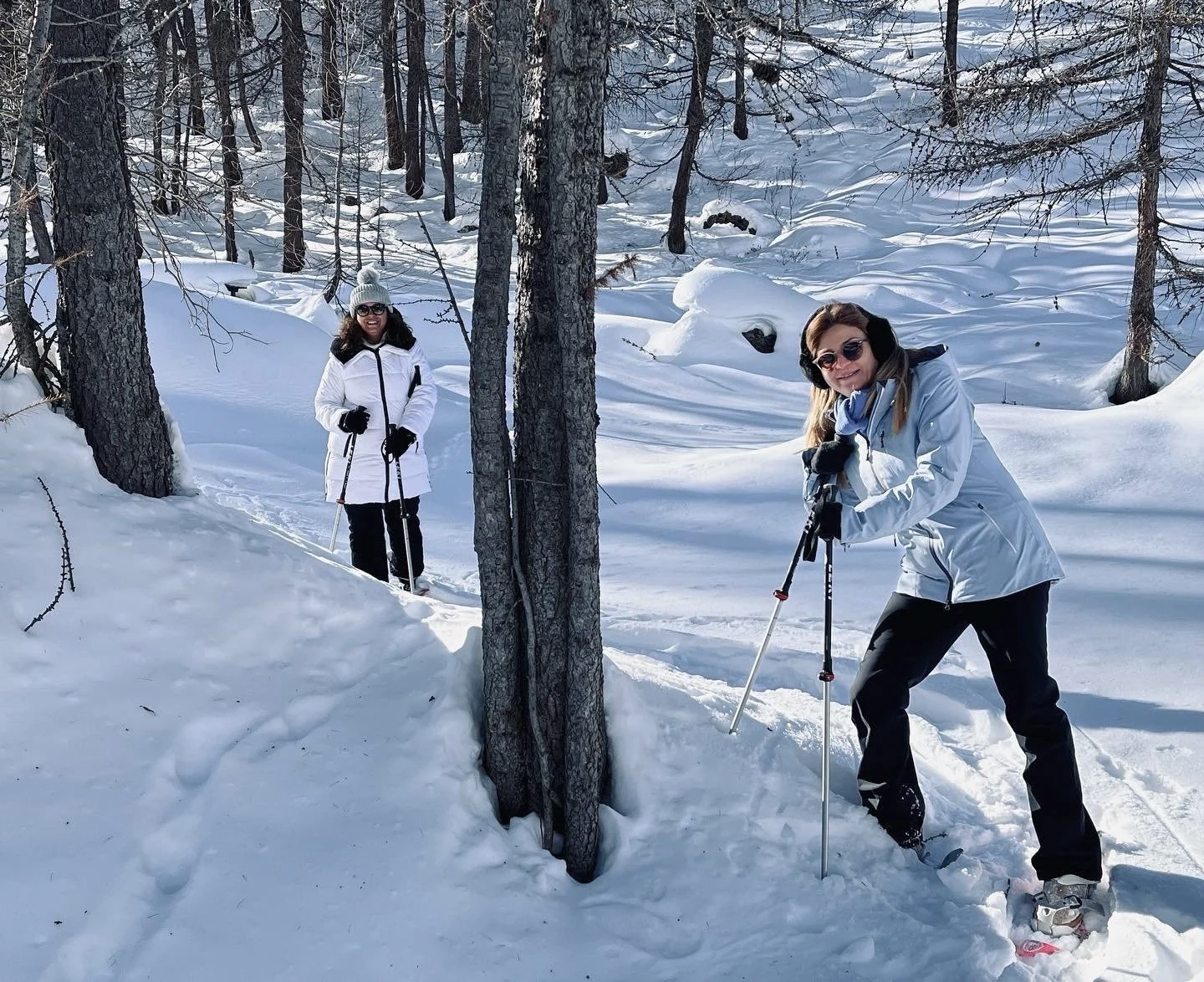 Two women snowshoeing in a snowy forest, smiling at the camera, wearing winter jackets, gloves, hats, and sunglasses.