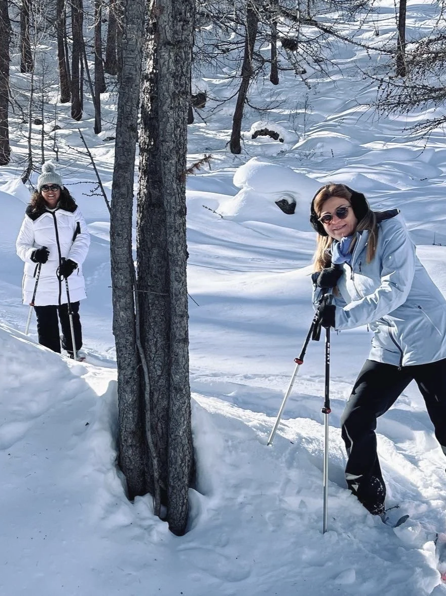 Two women wearing winter clothes and sunglasses are snowshoeing in a snow-covered forest with tall trees and snow drifts.