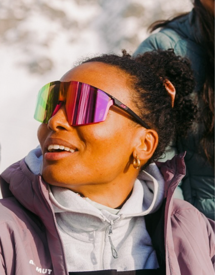 Woman wearing reflective ski goggles, gray hoodie, and purple jacket outdoors in a snowy setting.