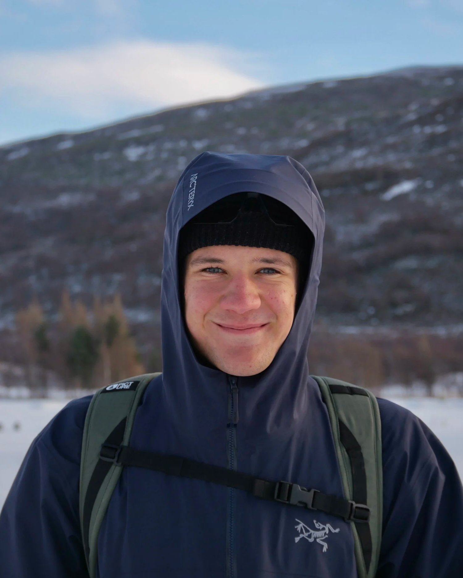 Young man outdoors in winter, wearing a dark jacket, black beanie, and backpack, smiling at camera with snowy mountain and trees in background.