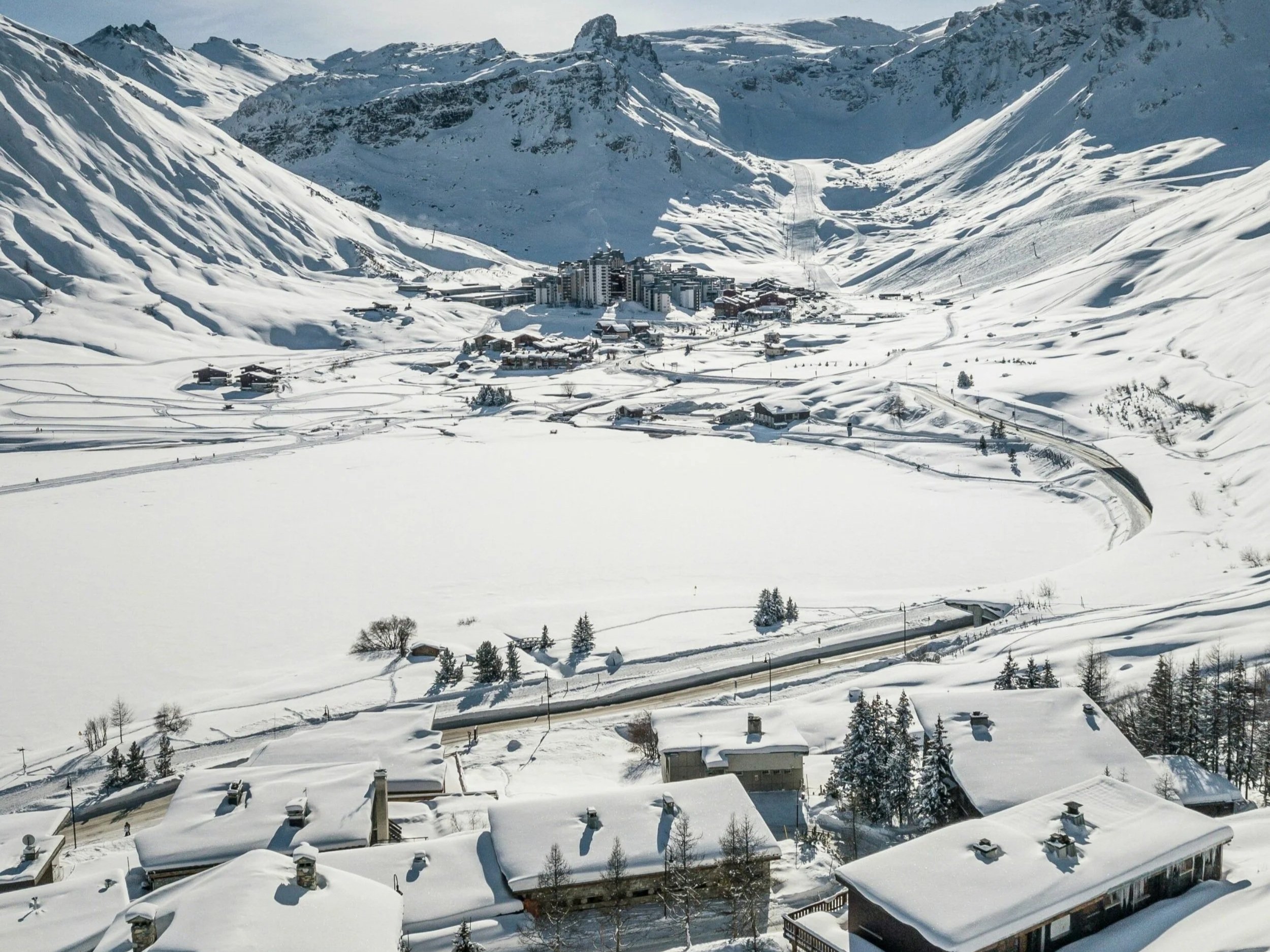 A snowy mountain landscape with a small village featuring buildings covered in snow, surrounded by snowed-in trees and winding roads, with larger mountains in the background.