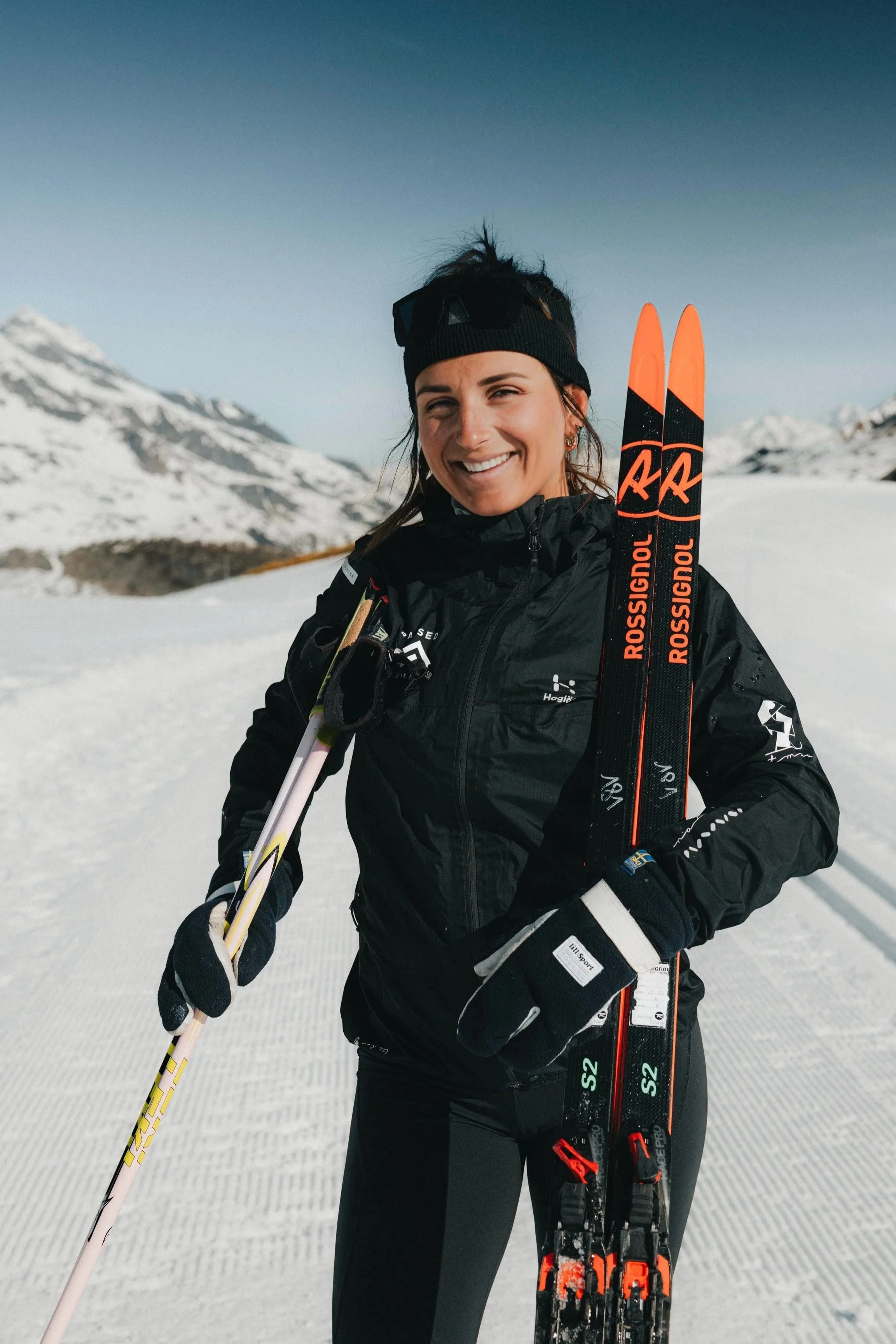 A woman in black winter sports gear holding skis and poles on a snowy mountain with clear sky and snow-covered peaks in the background.