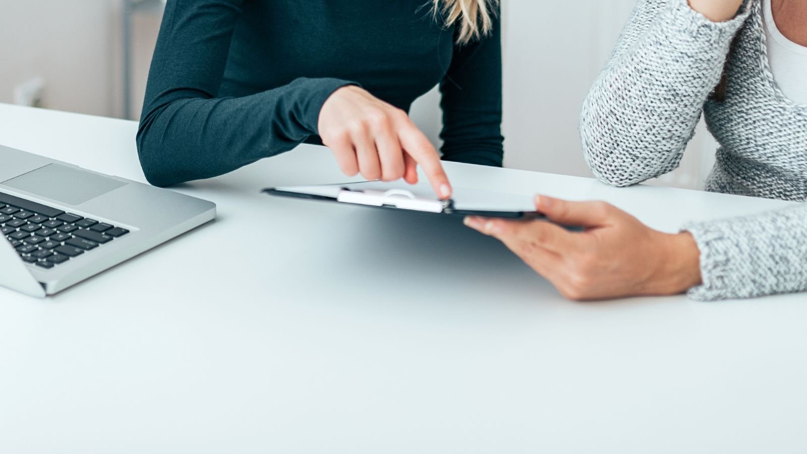 two women at a table, one holding a clipboard, the other pointing to something on it.
