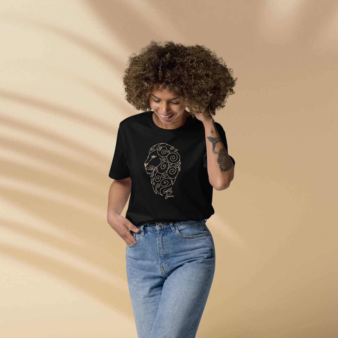 A woman with curly hair smiling and looking down, wearing a black t-shirt with a gold lion graphic and light blue jeans, standing against a beige background with soft shadow patterns.