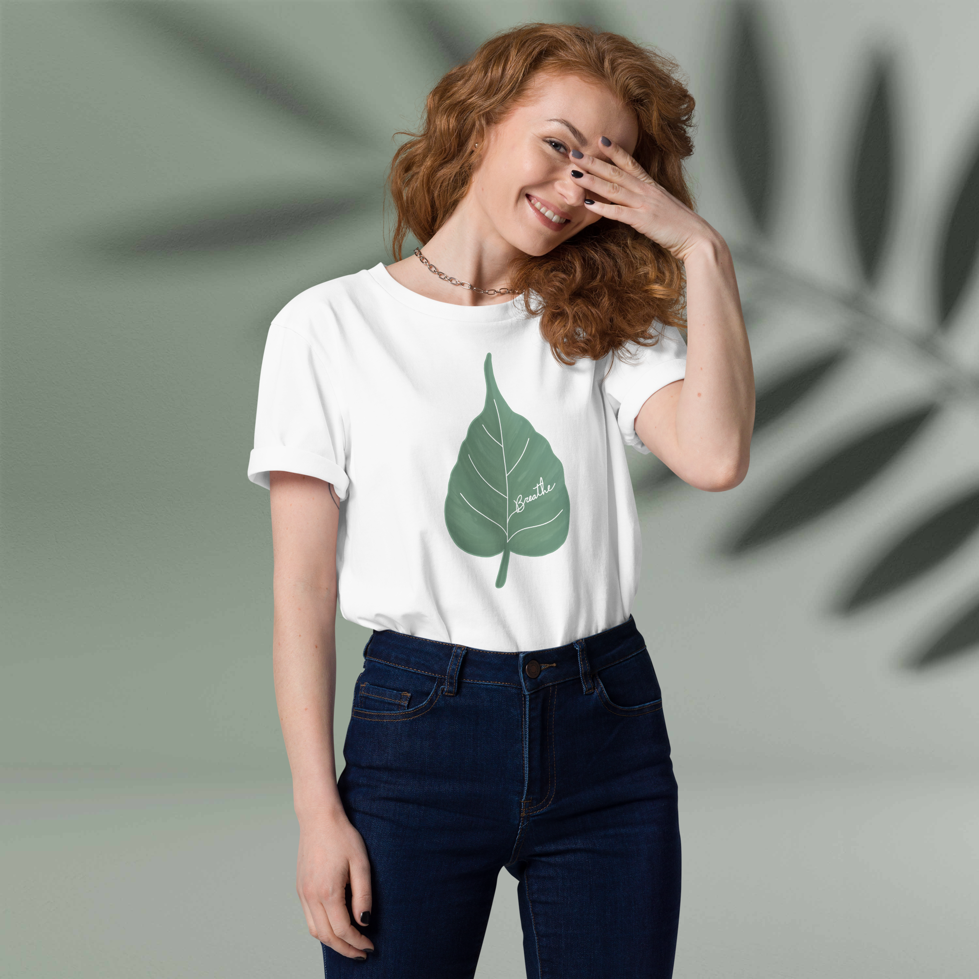 A woman with curly red hair smiling and covering her right eye with her hand, wearing a white T-shirt with a green leaf graphic and dark blue jeans.
