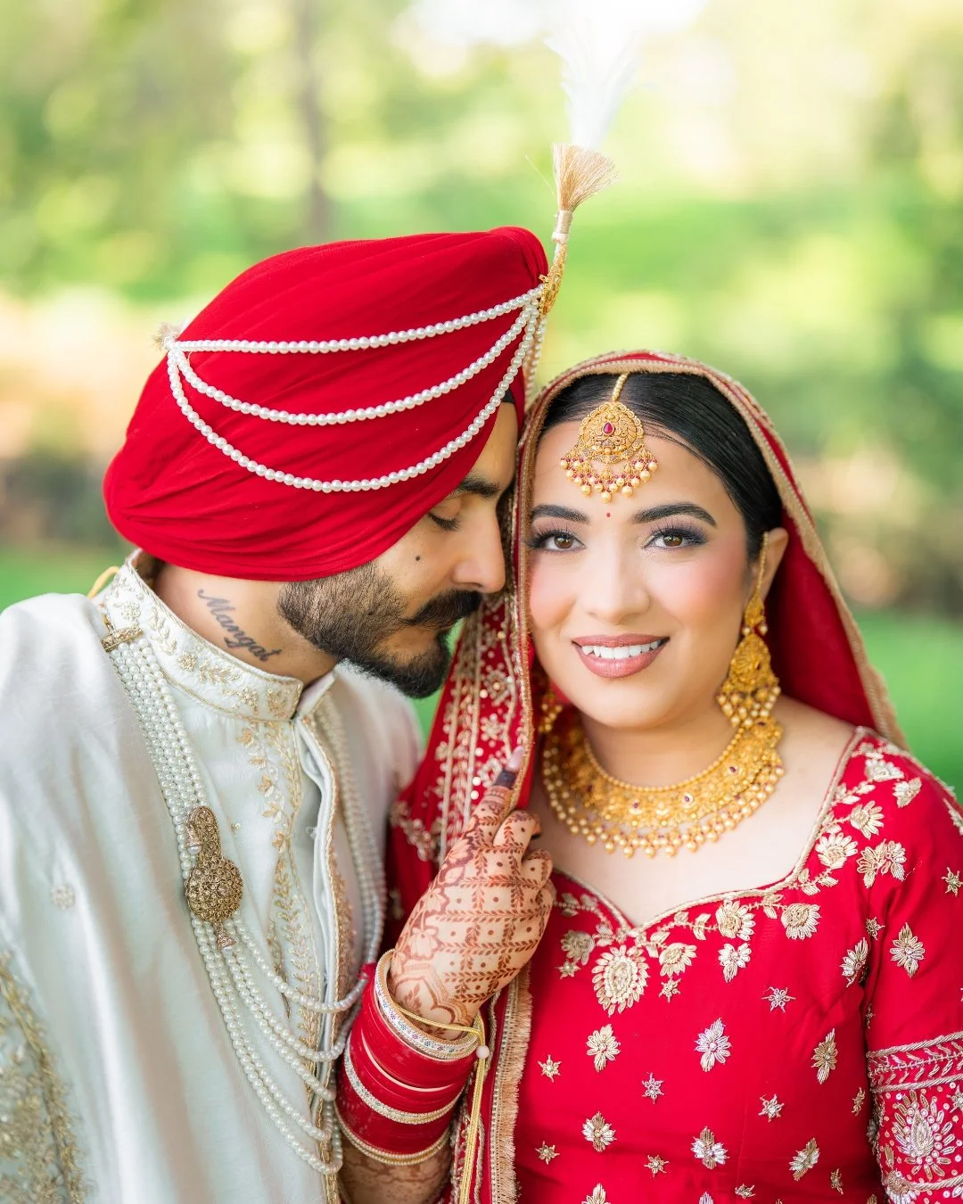 Indian bride and groom in traditional wedding attire outdoors with greenery in the background.