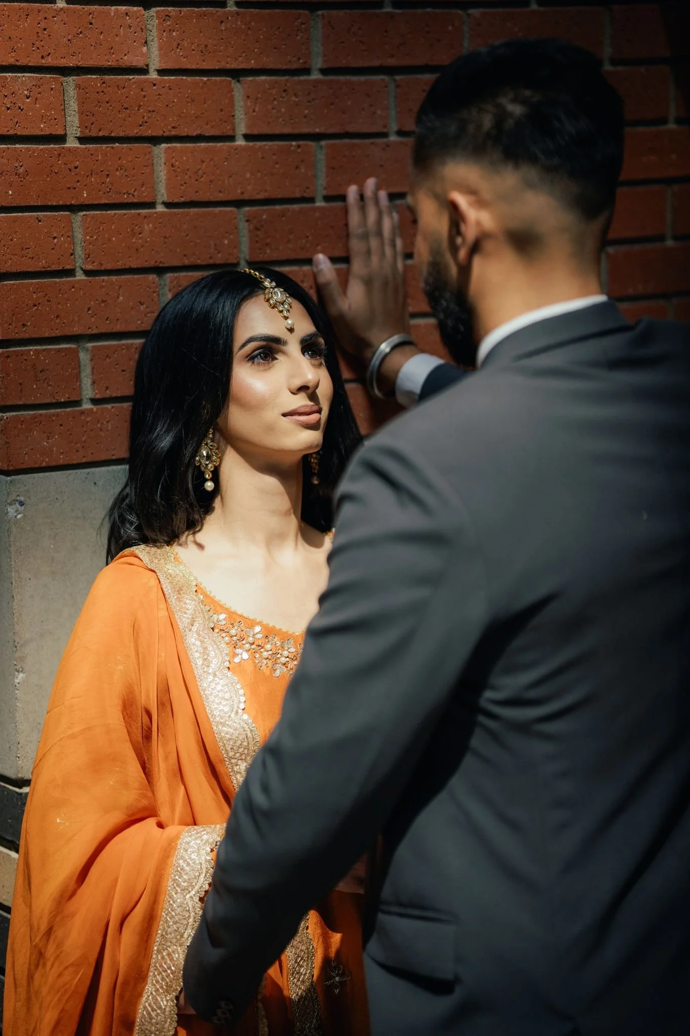 A woman dressed in traditional Indian attire and jewelry stands against a brick wall, while a man in a suit faces her with his hand on the wall.