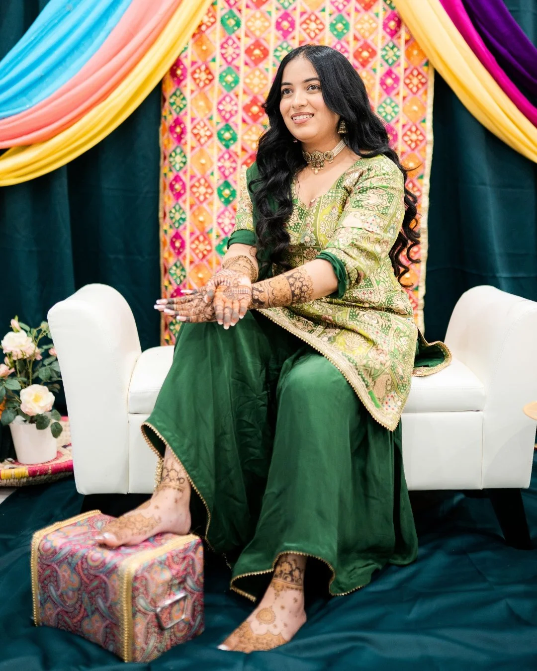 A woman dressed in a traditional green and gold Indian outfit, sitting on a white sofa with her hands decorated with henna, surrounded by colorful drapes and floral decorations, at a festive event.