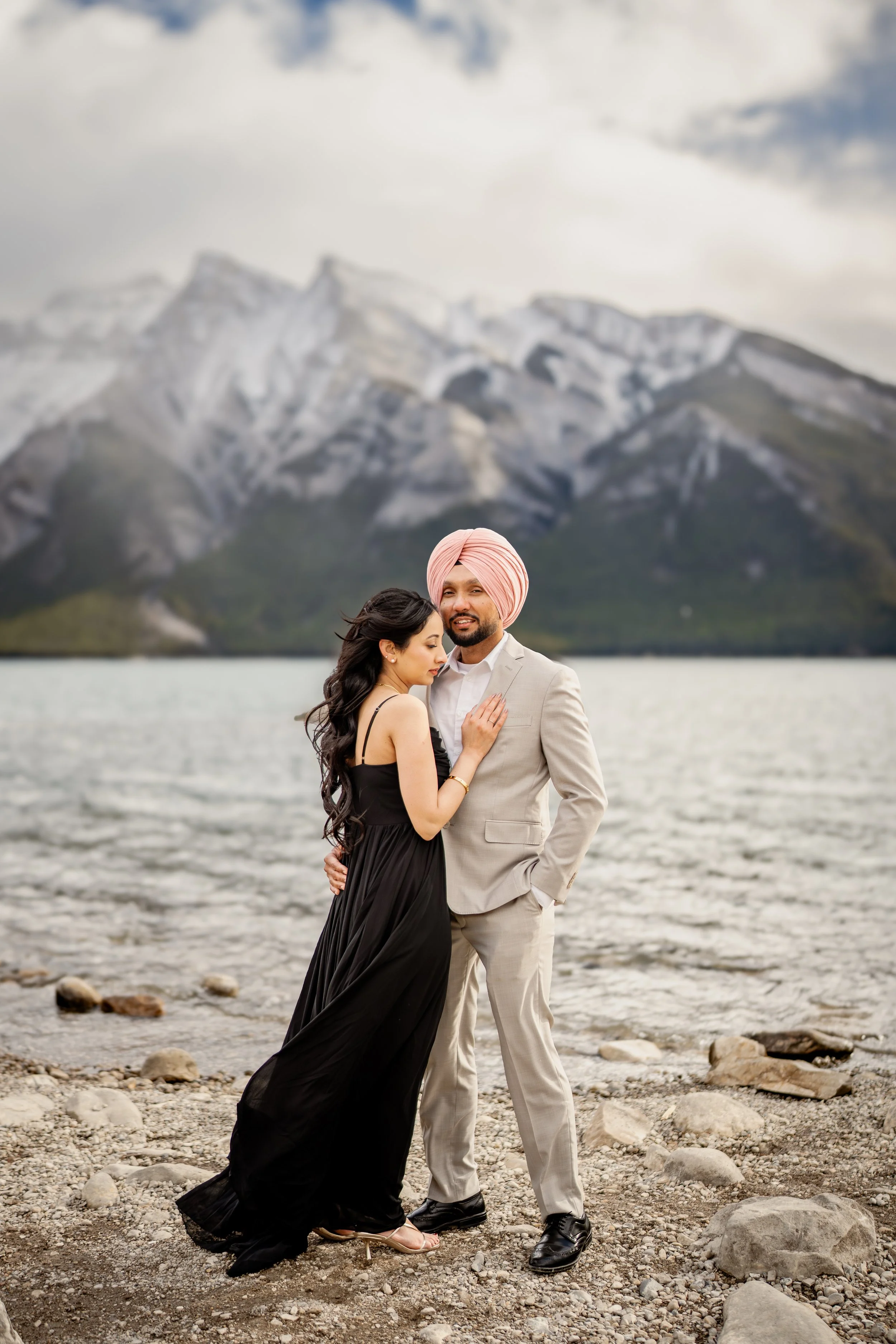 A couple standing on a rocky lakeshore with mountains in the background. The woman is in a black dress, and the man is in a light suit and pink turban.
