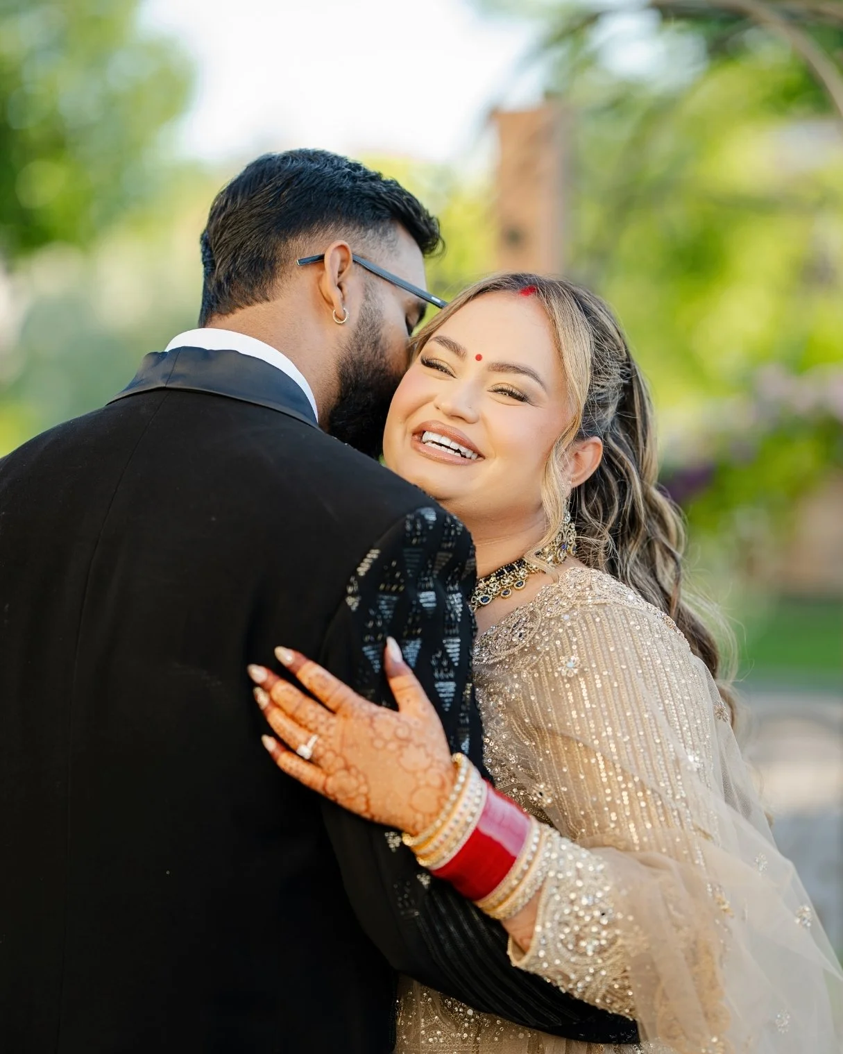 A married couple sharing a joyful hug outdoors, dressed in traditional wedding attire, with the bride smiling and the groom leaning in for a kiss.