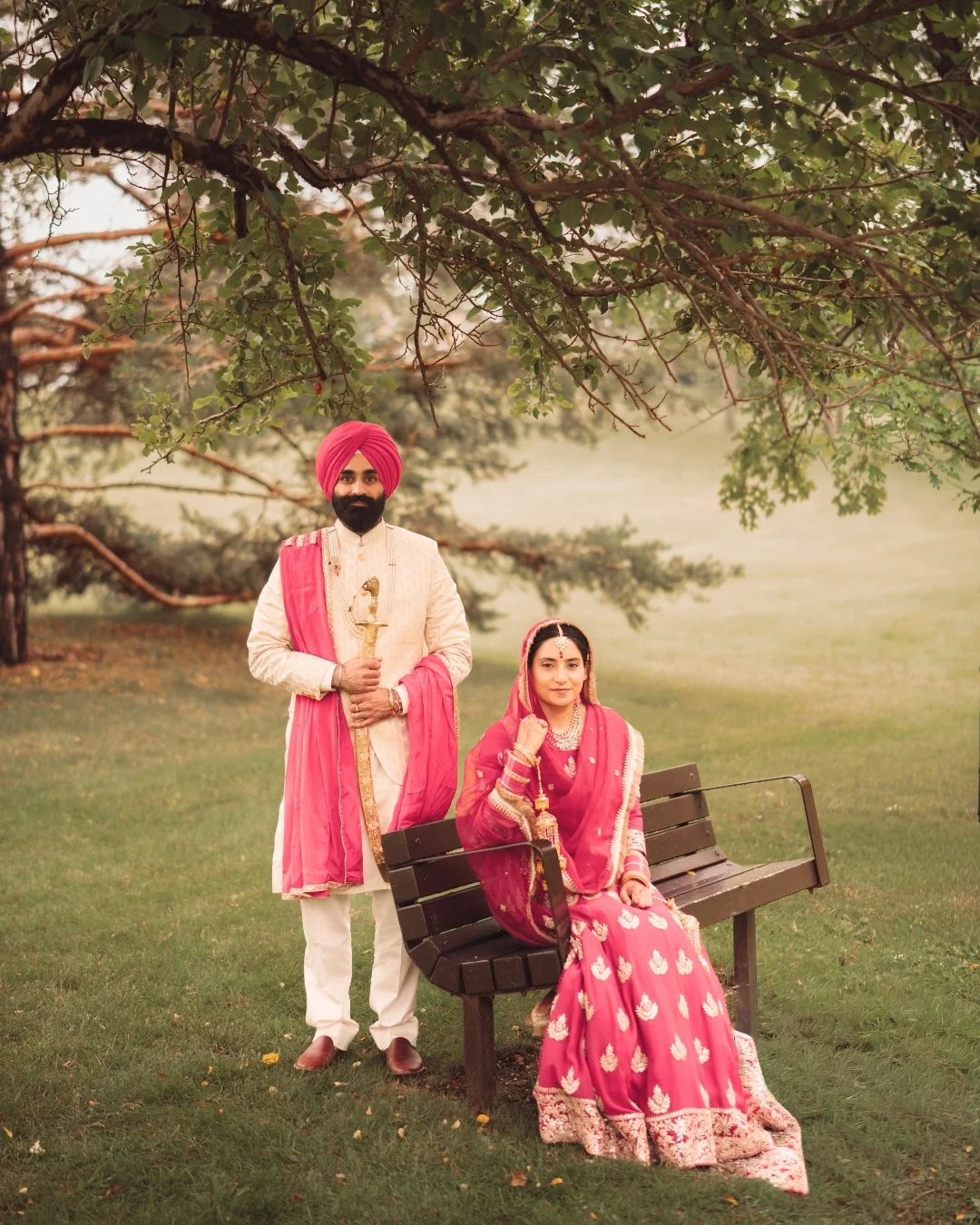A couple dressed in traditional Indian attire, the man wearing a pink turban and cream sherwani with a pink shawl, holding a staff, and the woman seated on a park bench wearing a pink and gold embroidered sari and jewelry, in a lush green park with t