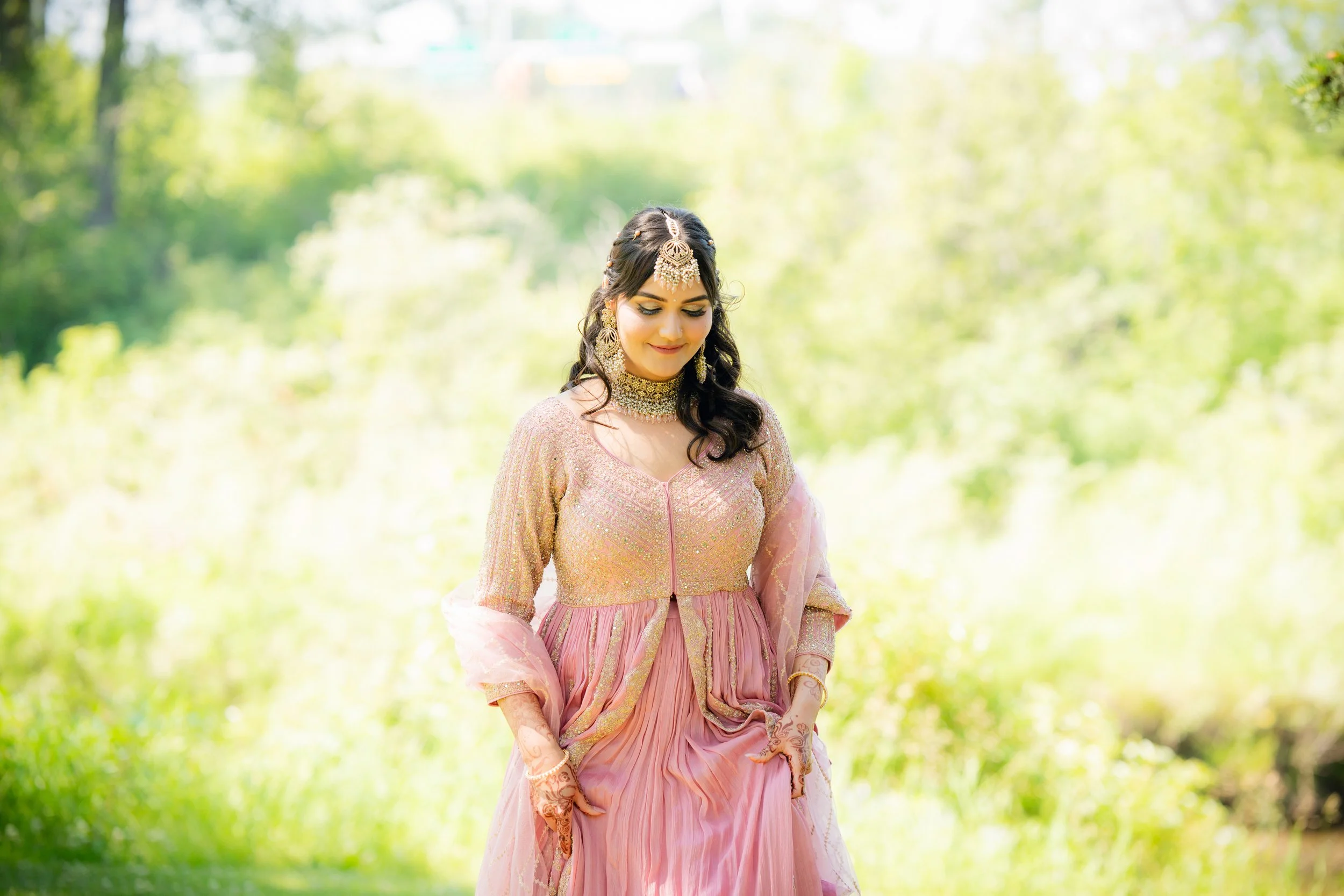 A woman dressed in traditional Indian attire standing outdoors surrounded by green foliage, looking down with a gentle smile, wearing jewelry including earrings, a necklace, and a headpiece.